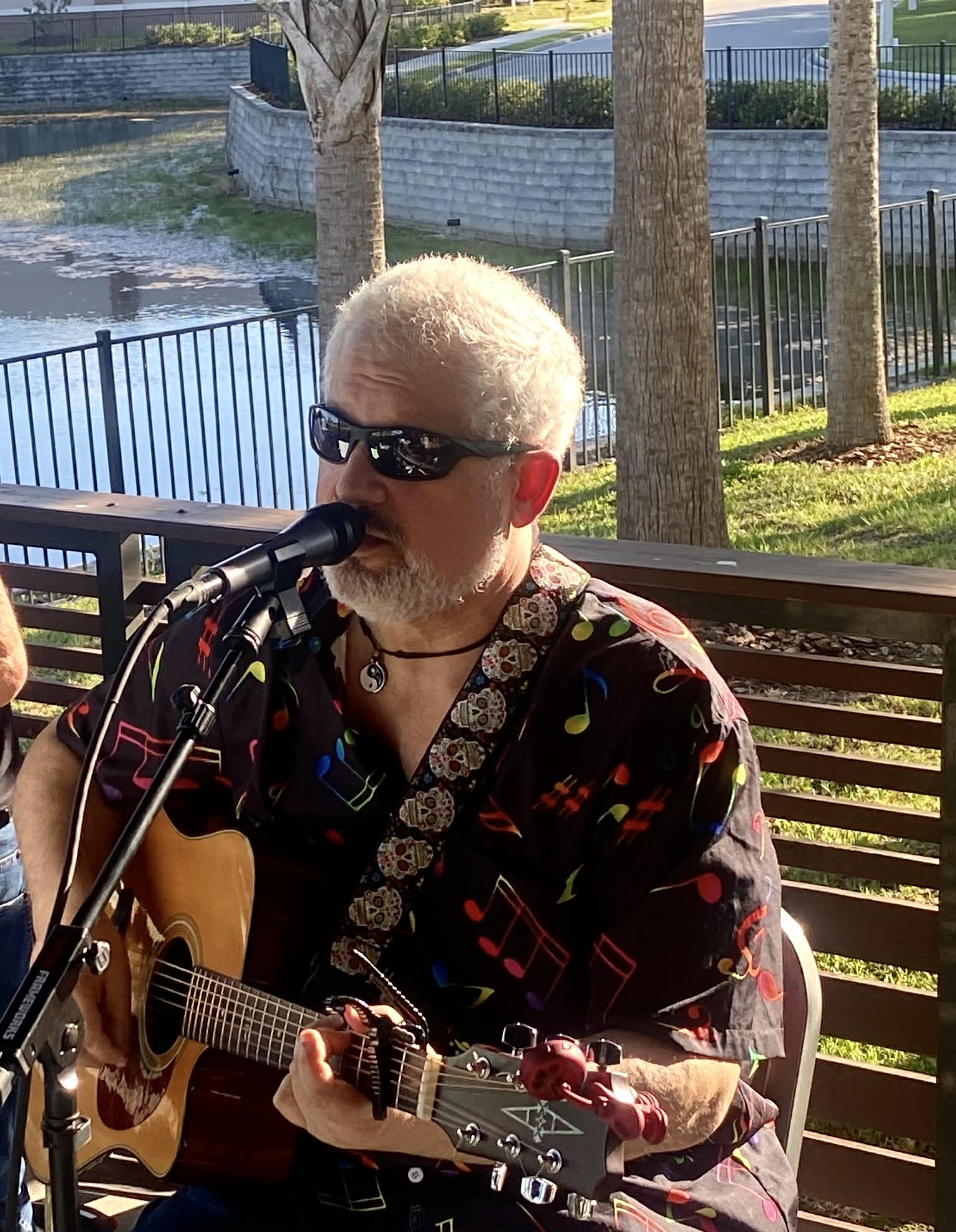 Person with white hair and sunglasses playing guitar and singing outdoors near trees and a pond.