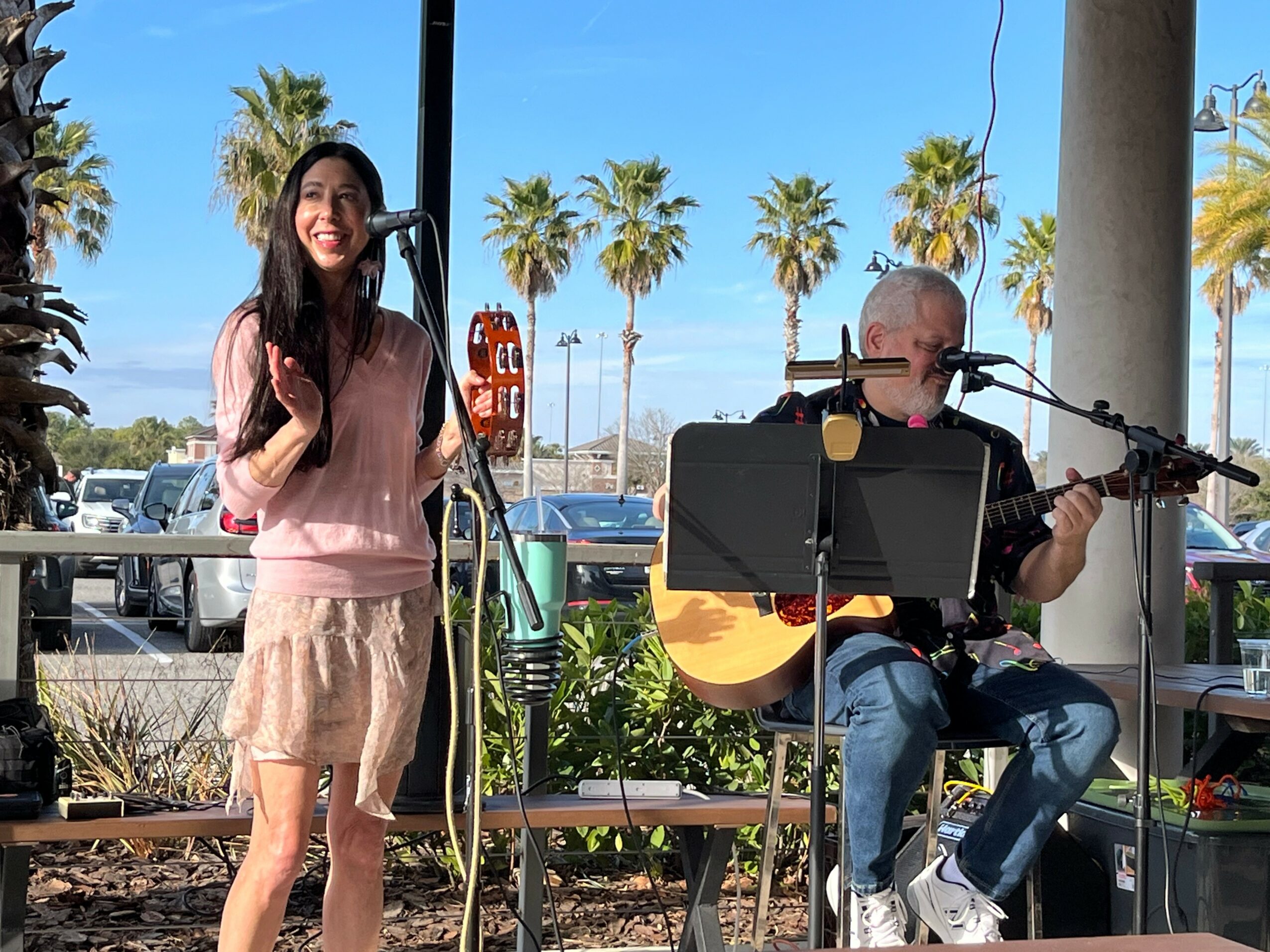 A woman singing with a microphone and a man playing guitar outdoors near palm trees, with a blue sky.