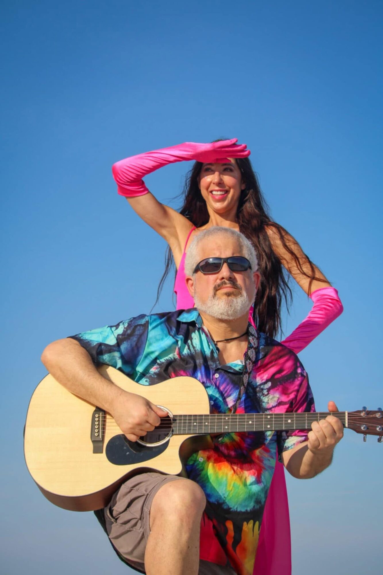 A man with sunglasses playing guitar, a woman in pink gloves smiling behind him, against a clear blue sky.
