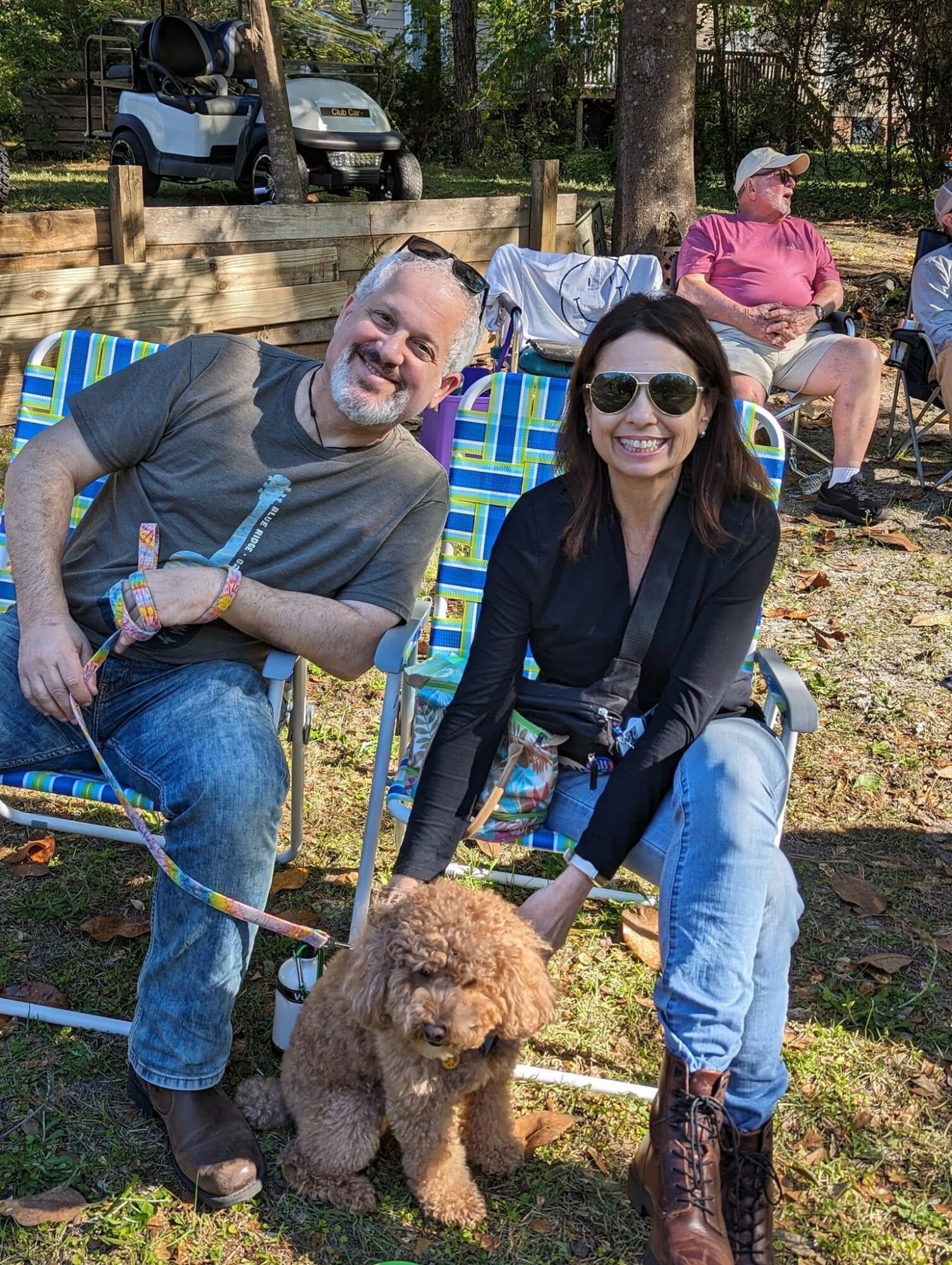 Two people sitting outdoors with a brown poodle, smiling, wearing sunglasses, and casual clothing, in a park-like setting.