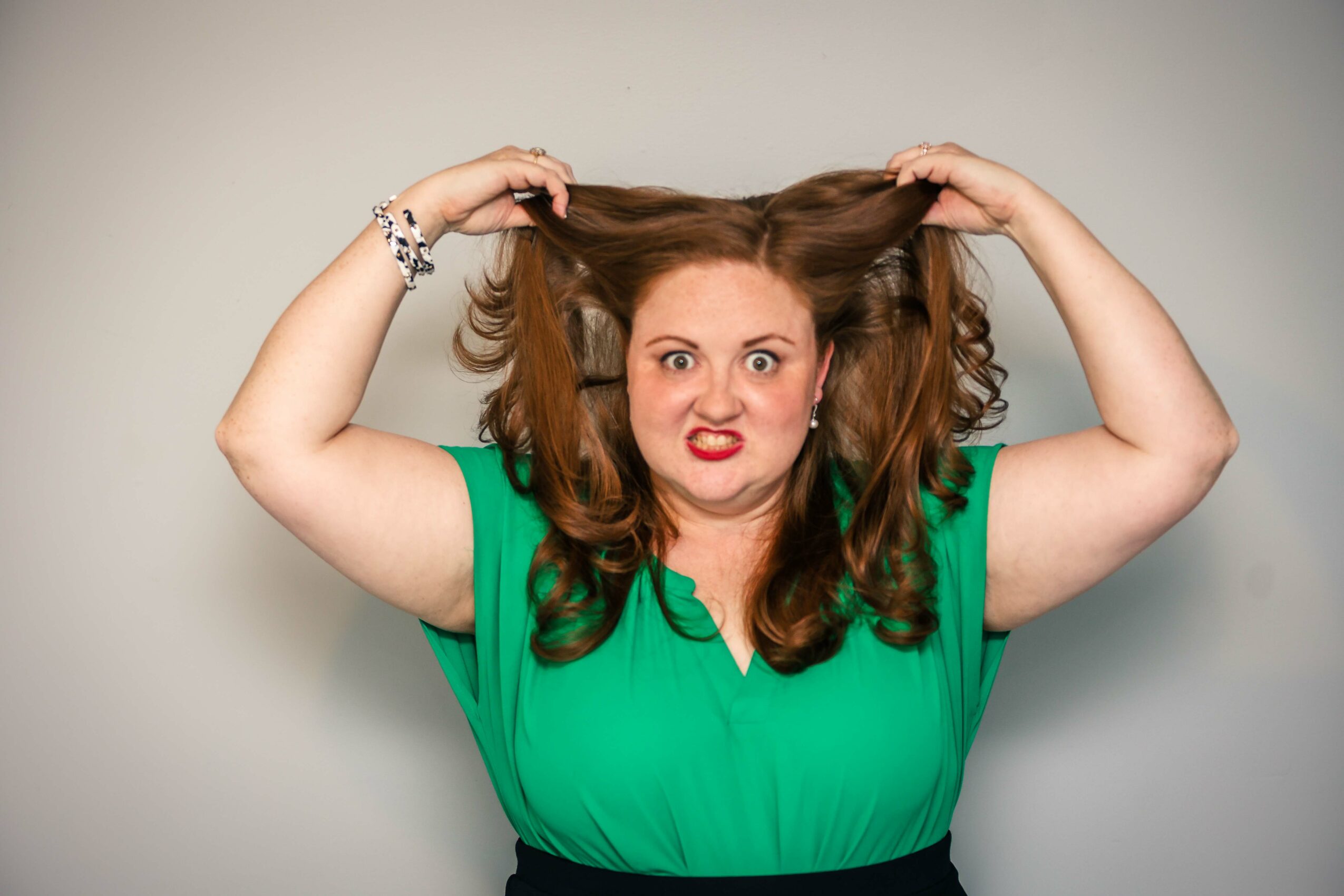 Woman with long brown hair pulling it up, wearing a green top, making a frustrated facial expression, against a plain background.