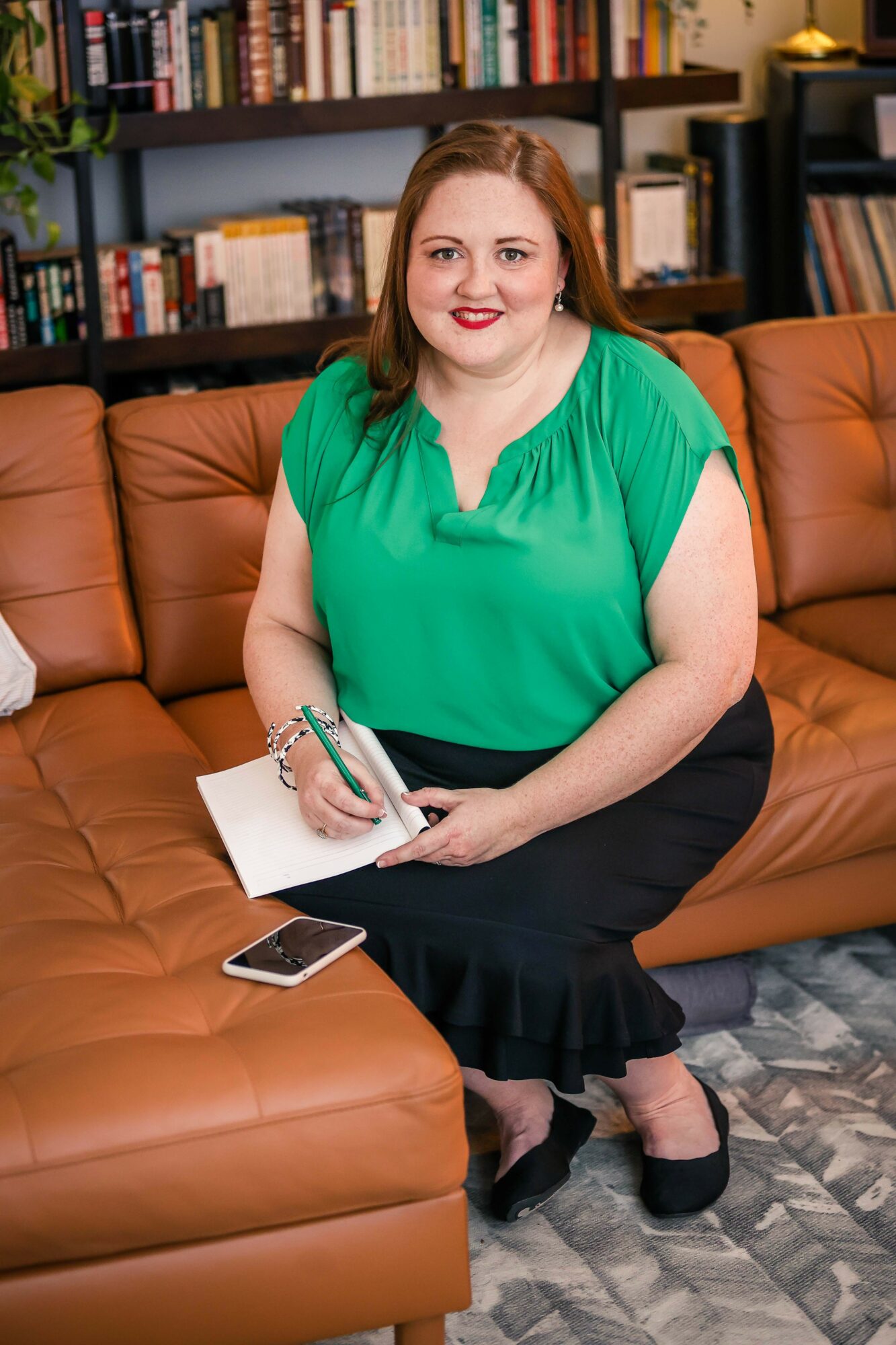 Woman sitting on a brown leather couch holding a notebook and pen, with a bookshelf in the background.