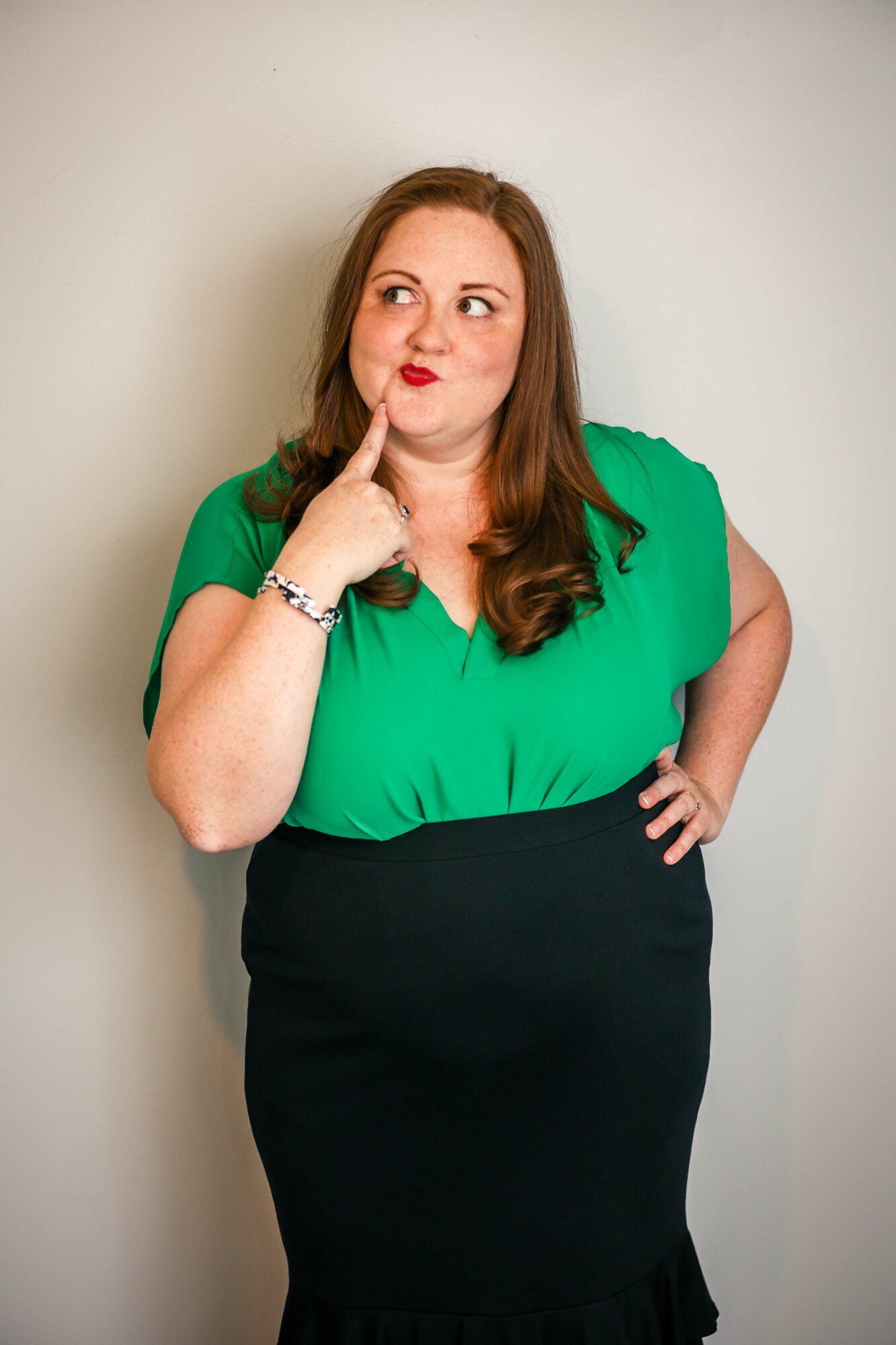 Woman with long brown hair in a green top and black skirt, touching her chin thoughtfully, standing against a plain background.