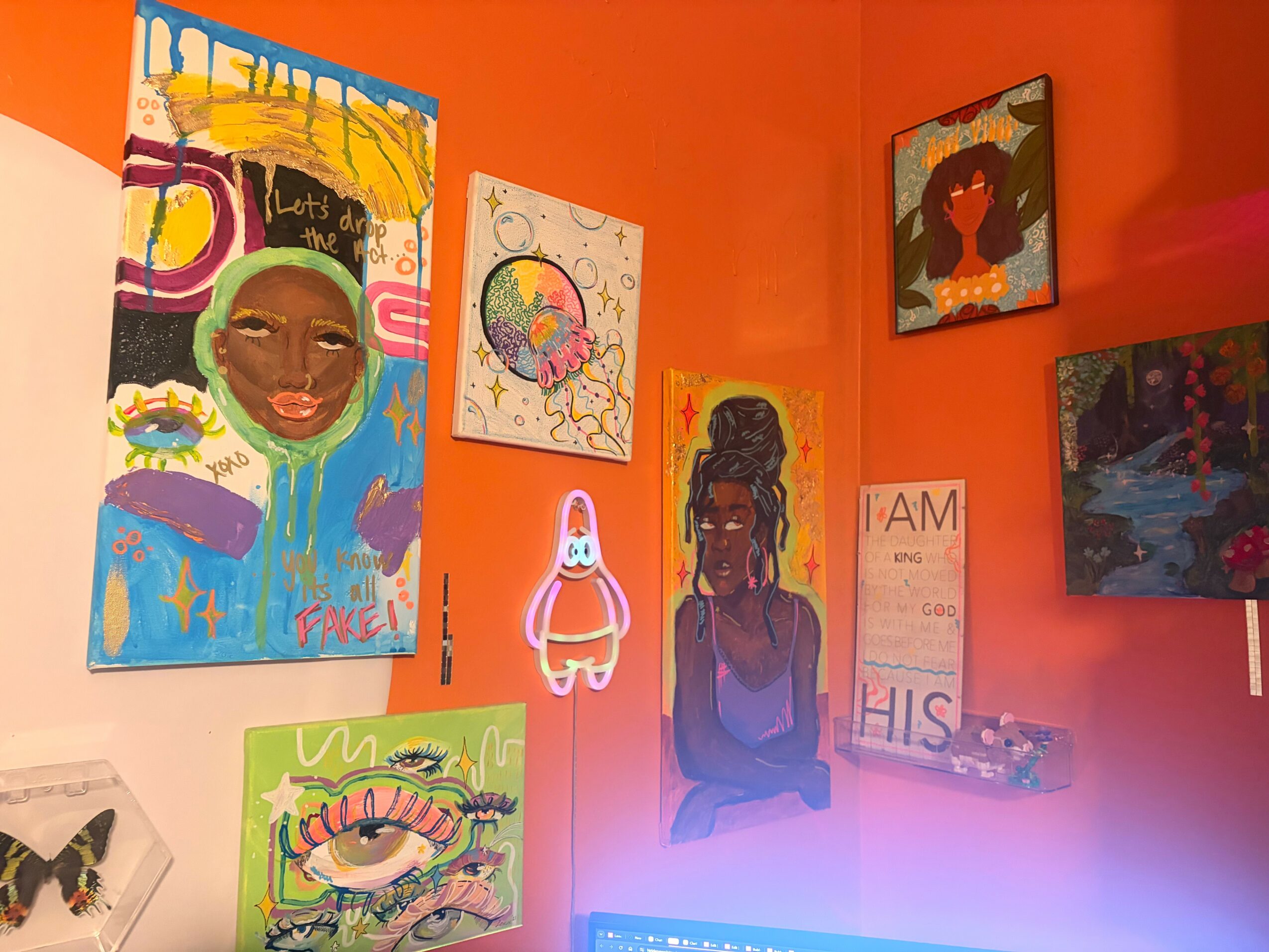 Gallery wall with colorful paintings and drawings on orange walls, including a face, abstract art, and a bunny figure.