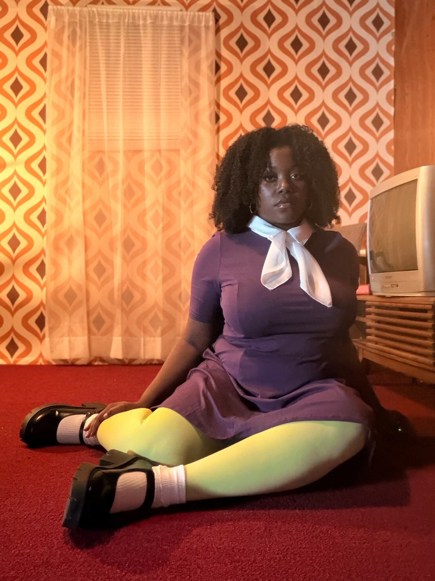 Young woman with curly hair sitting on a red carpet, wearing a purple dress, yellow tights, and black shoes, in a room with patterned curtains and a vintage TV.