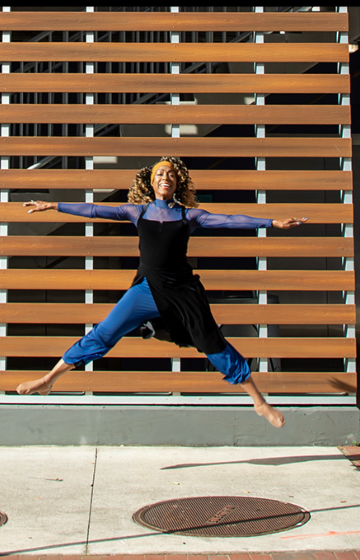 Person jumping with arms and legs outstretched in front of a wooden slat wall, smiling, outdoors.
