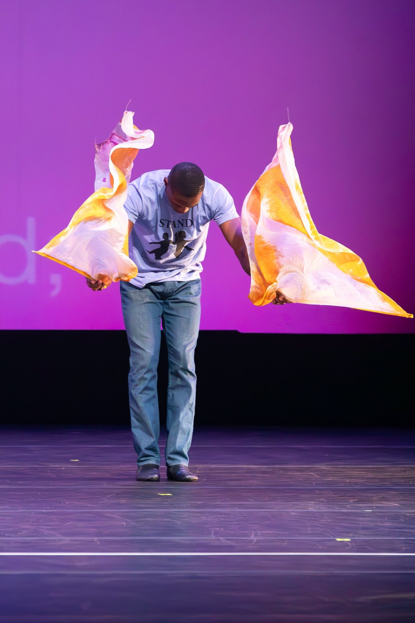 Person on stage with yellow and white fabric flowing, purple background, wooden floor, bowing head, arms extended downward.