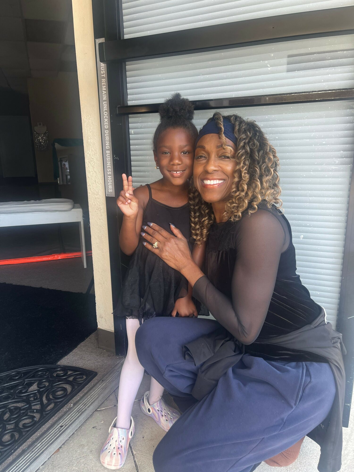 Two women sitting on a porch, one with curly hair and the other with straight hair, smiling and making peace signs.