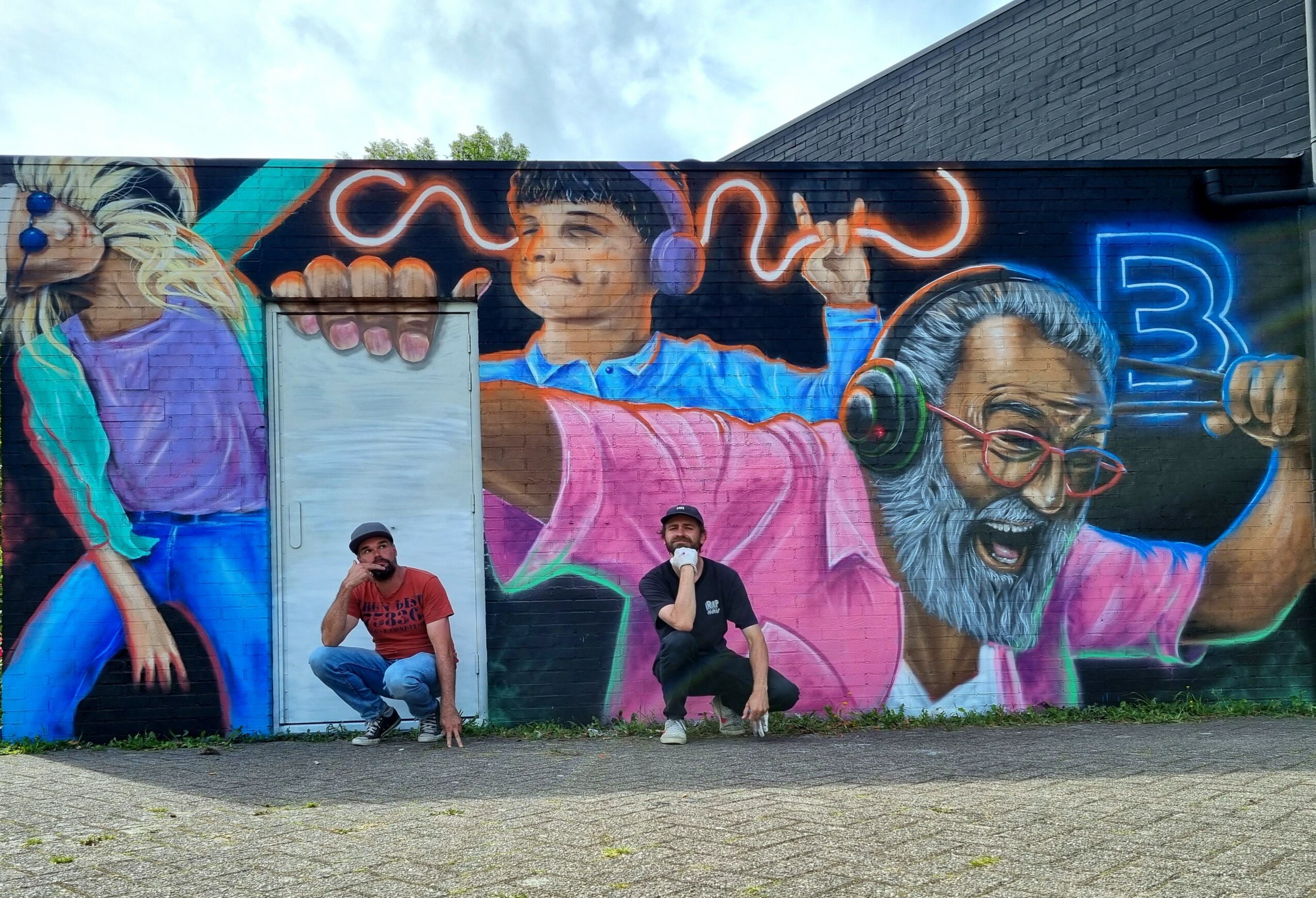 Colorful mural of three people, one with headphones, on a wall outdoors, with two people crouching in front, one holding a phone.