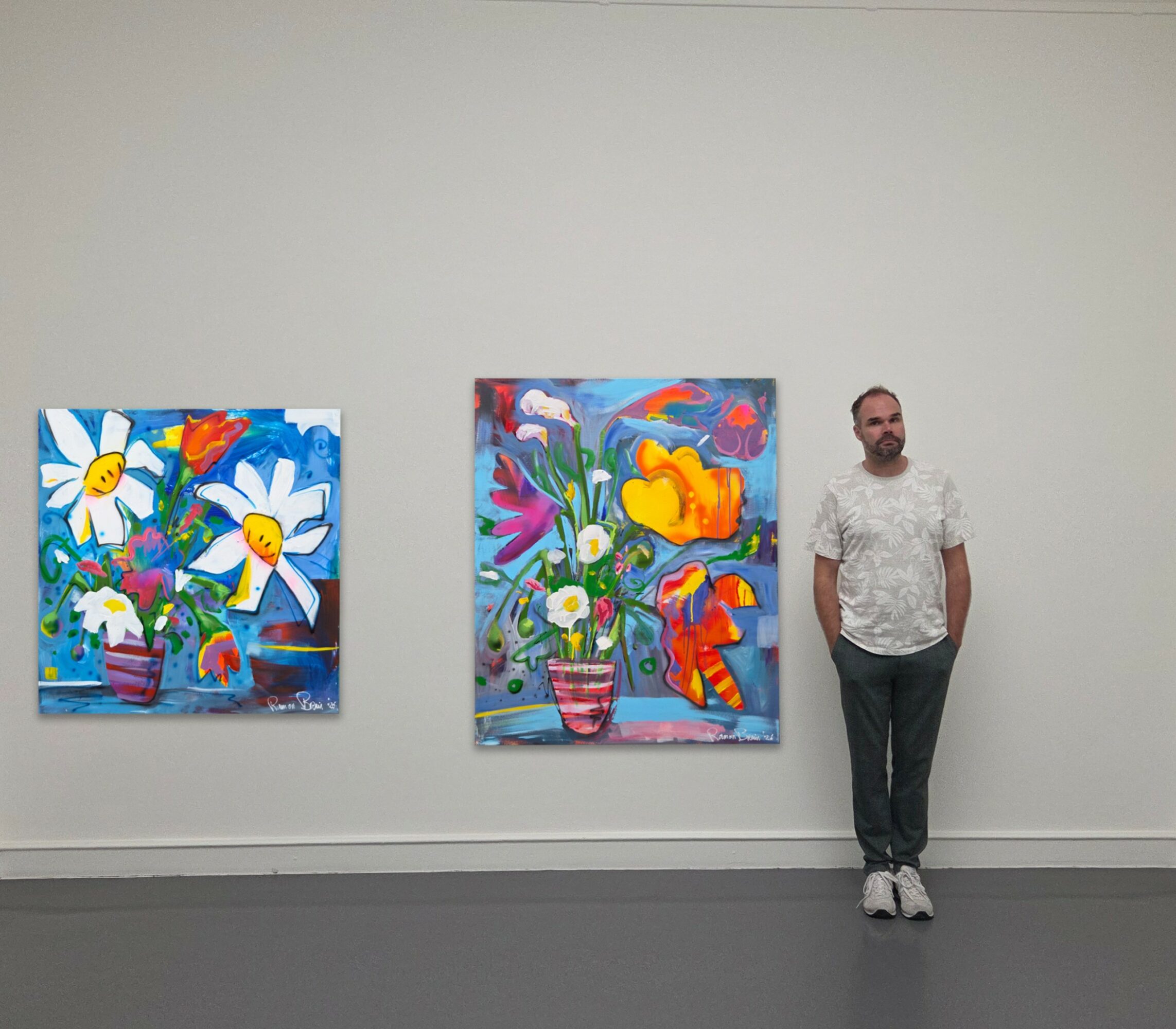 Man standing next to two colorful abstract paintings on a white gallery wall.