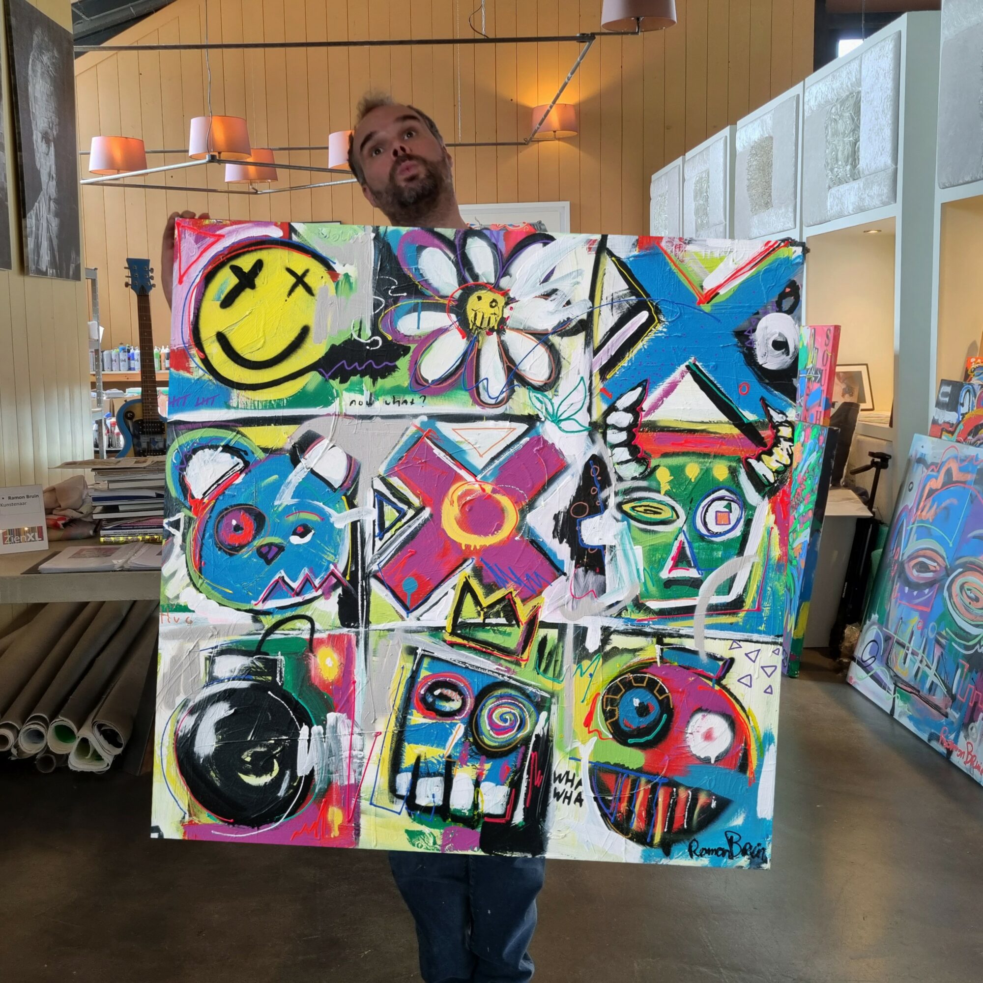 Person holding a colorful, abstract painting with various shapes and smiley faces in an indoor space.