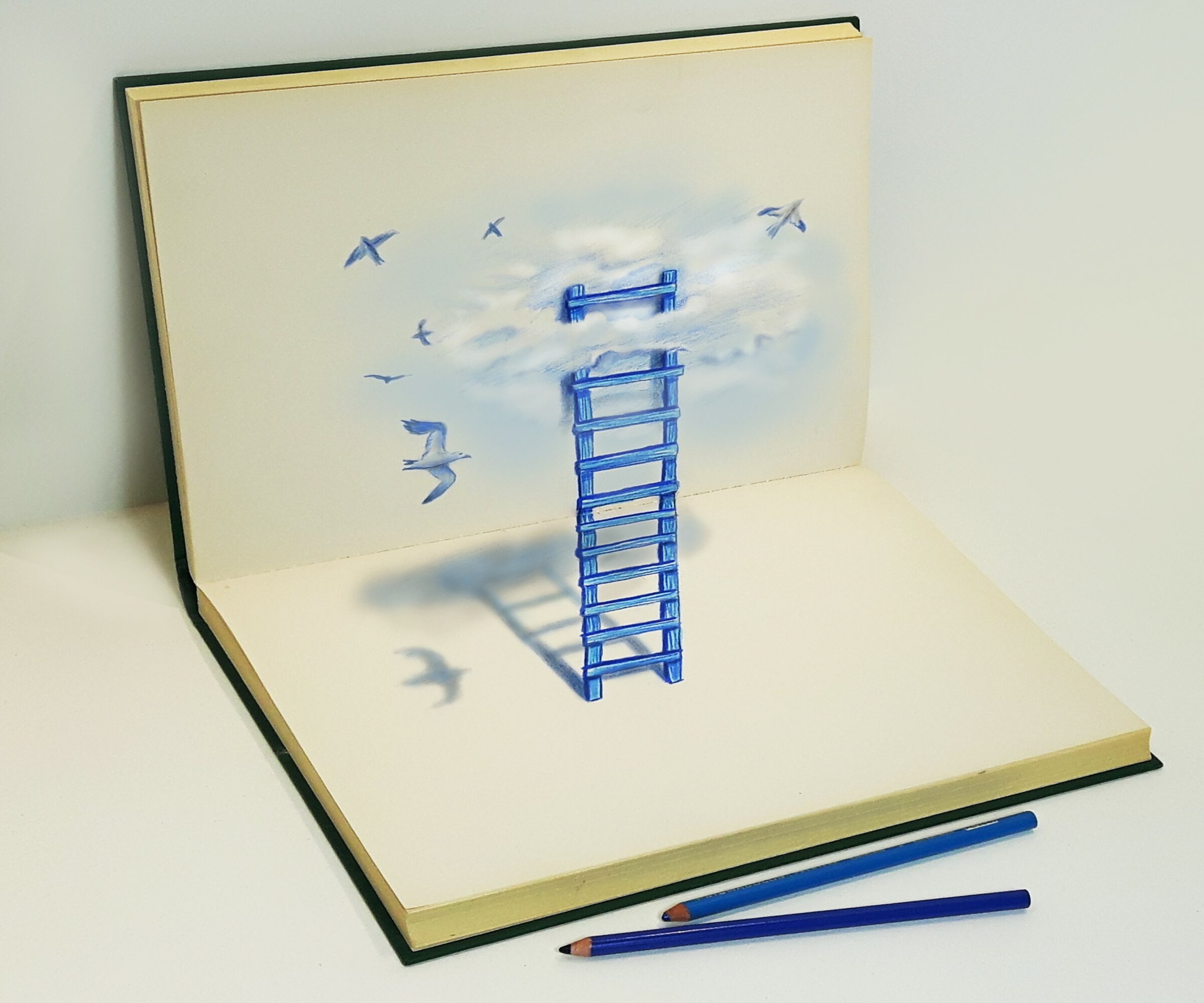 Open sketchbook with a blue ladder drawing leading to clouds and birds, two pens lying in front, on a white surface.