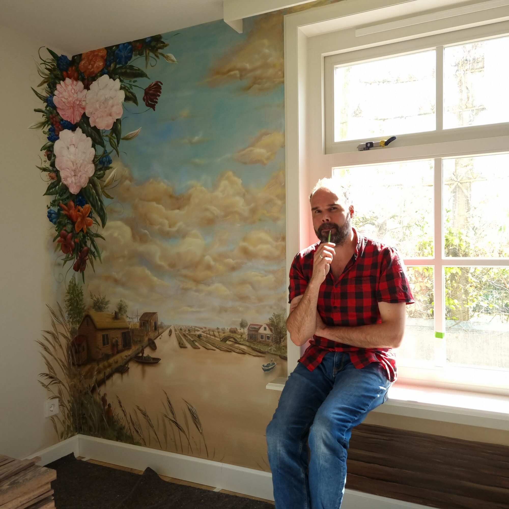 Man with glasses and beard sitting on window sill, wearing red plaid shirt and jeans, with a mural of a landscape behind him.