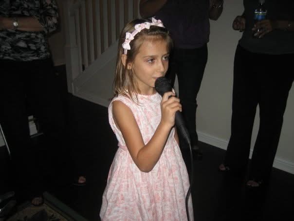 Young girl with a headband holding a microphone, standing indoors, with people in the background.