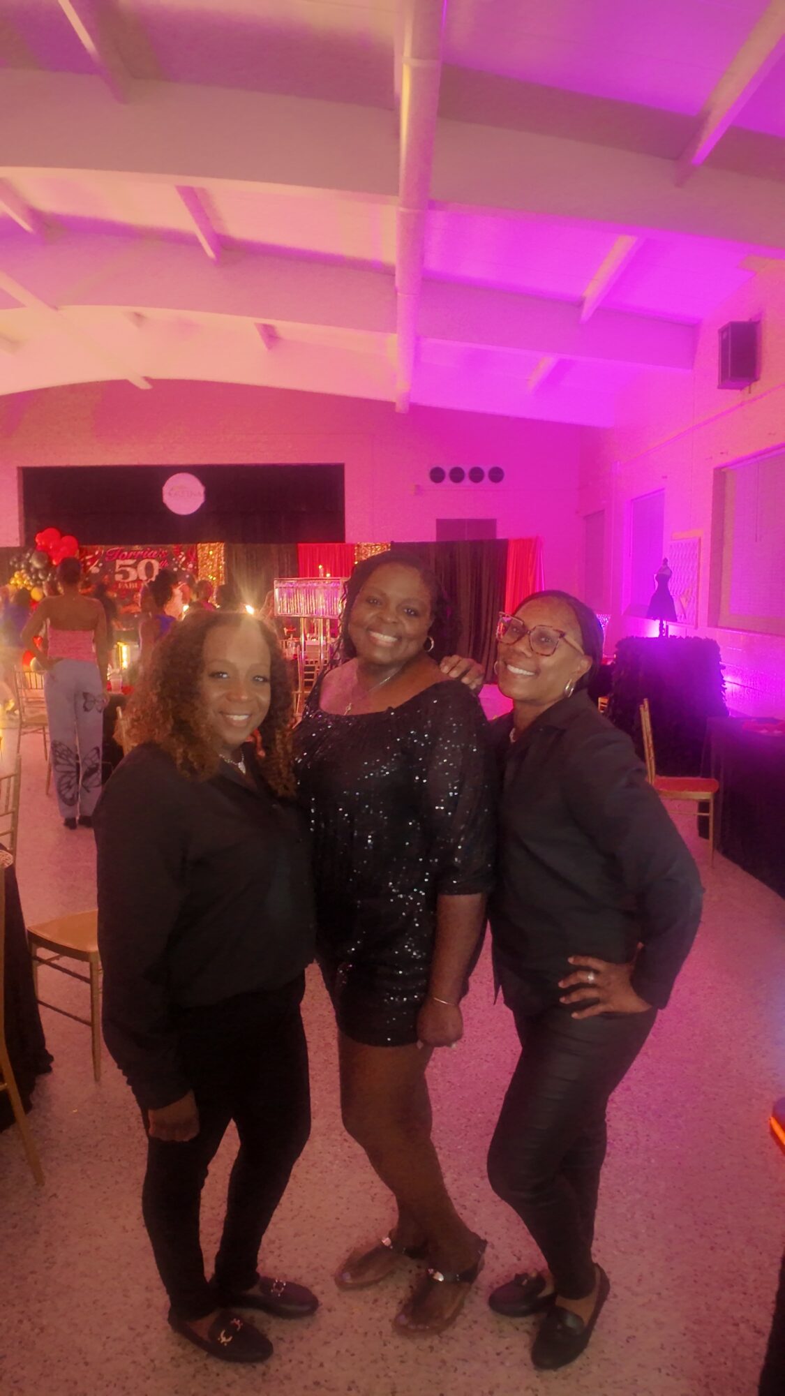 Three women standing together in a decorated indoor event space with pink and purple lighting, smiling at the camera.