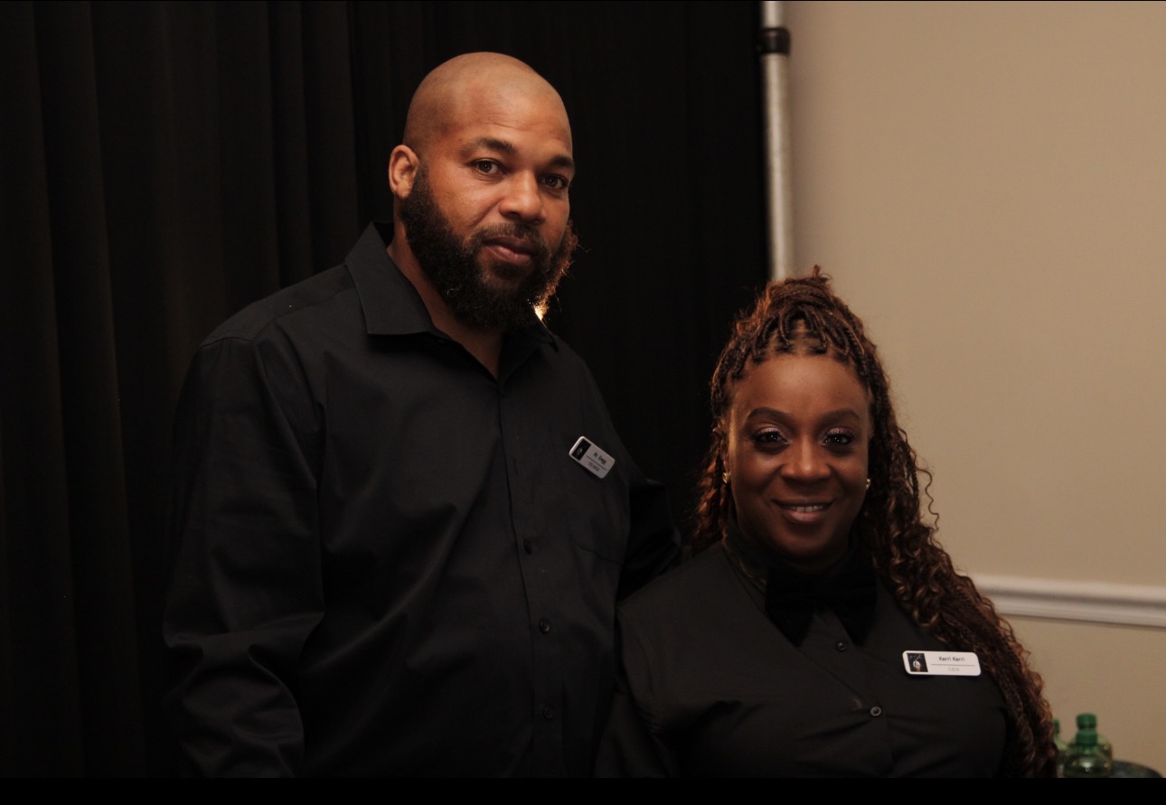 Two people standing indoors, both wearing black shirts, smiling, with a dark curtain and beige wall behind them.