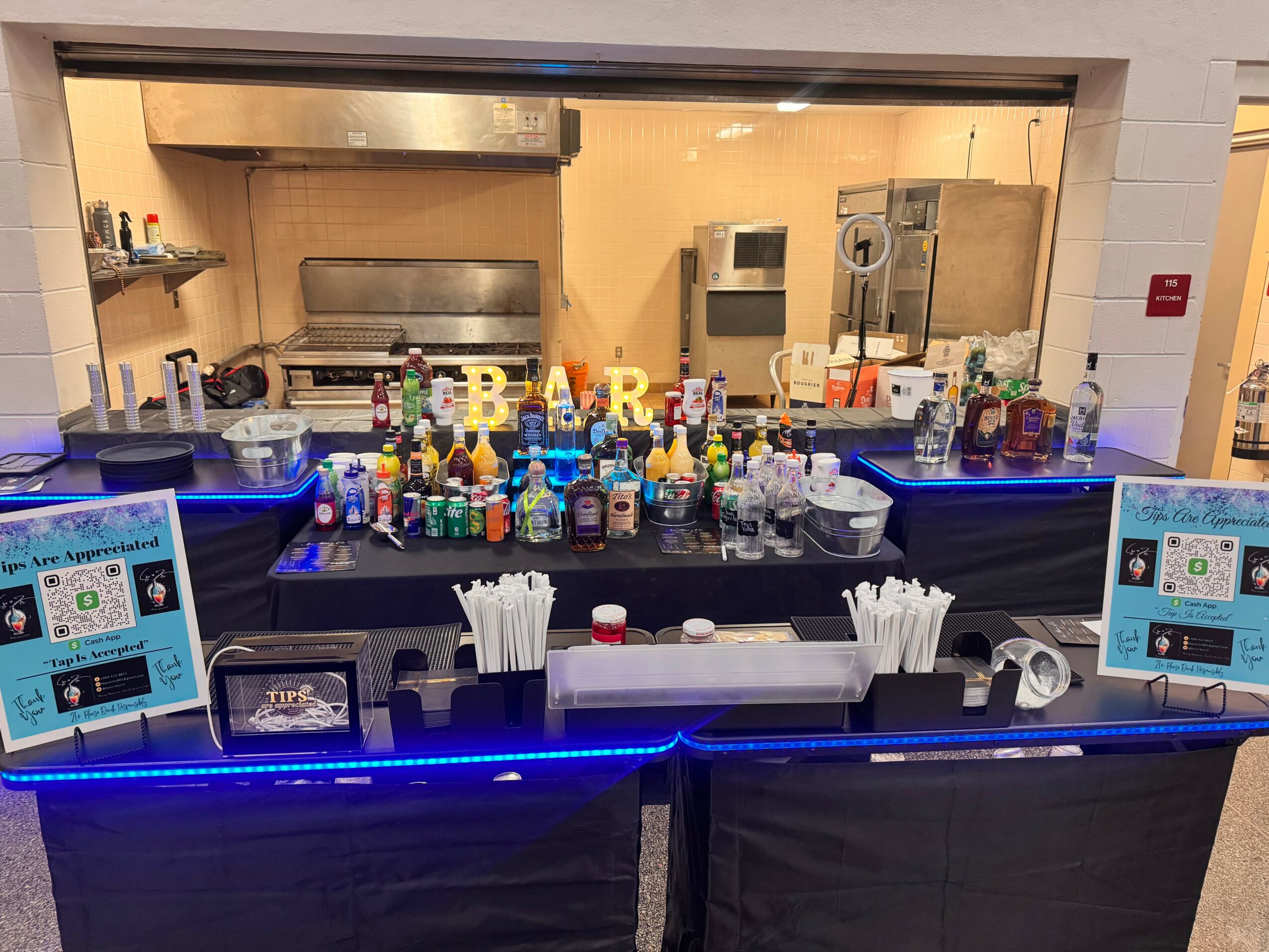 Bar counter with bottles, glasses, and two digital screens, viewed through a window from the kitchen area behind the bar.