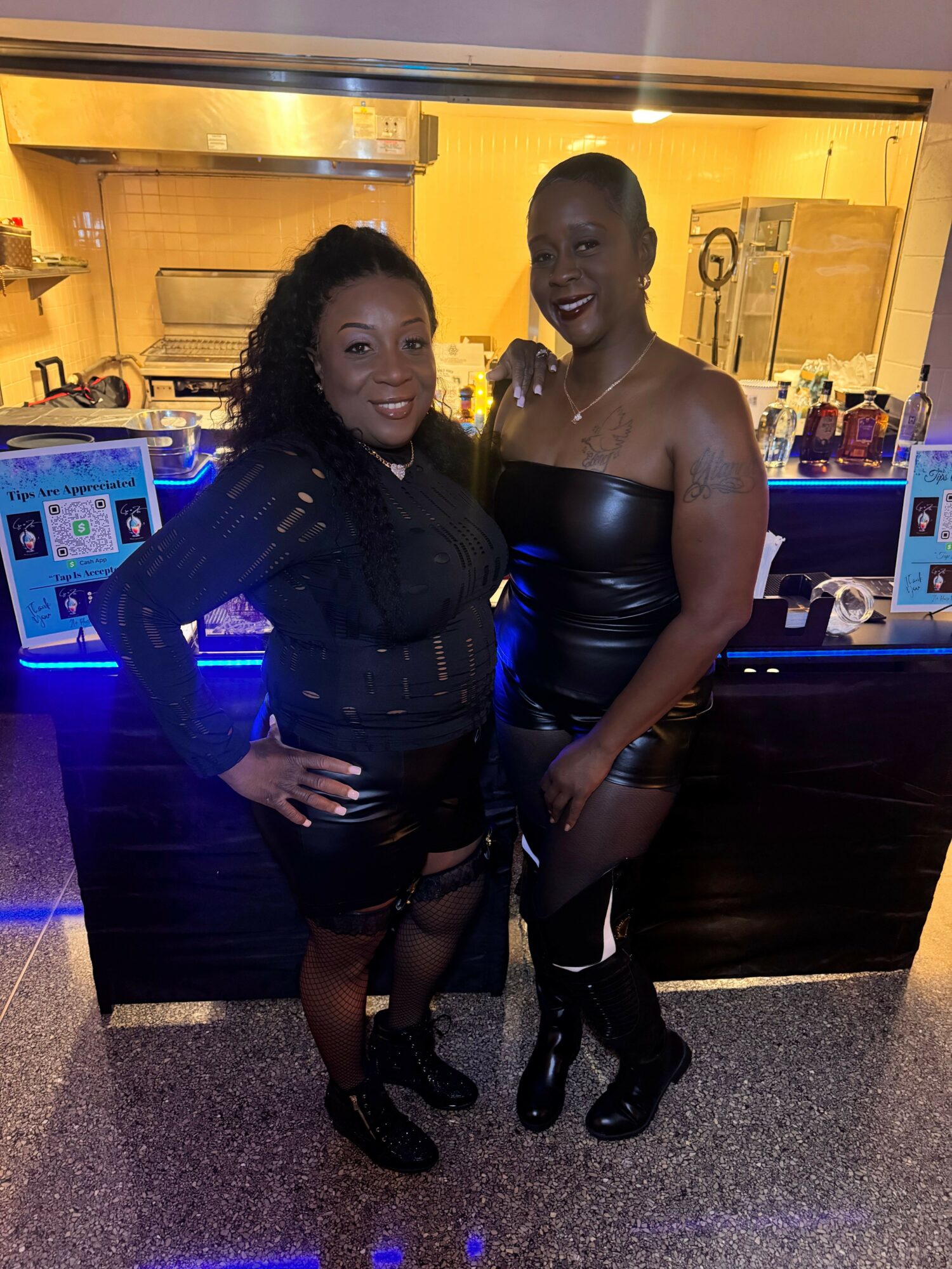 Two women pose together in a nightclub, one in a black dress and the other in a black outfit, with a bar in the background.