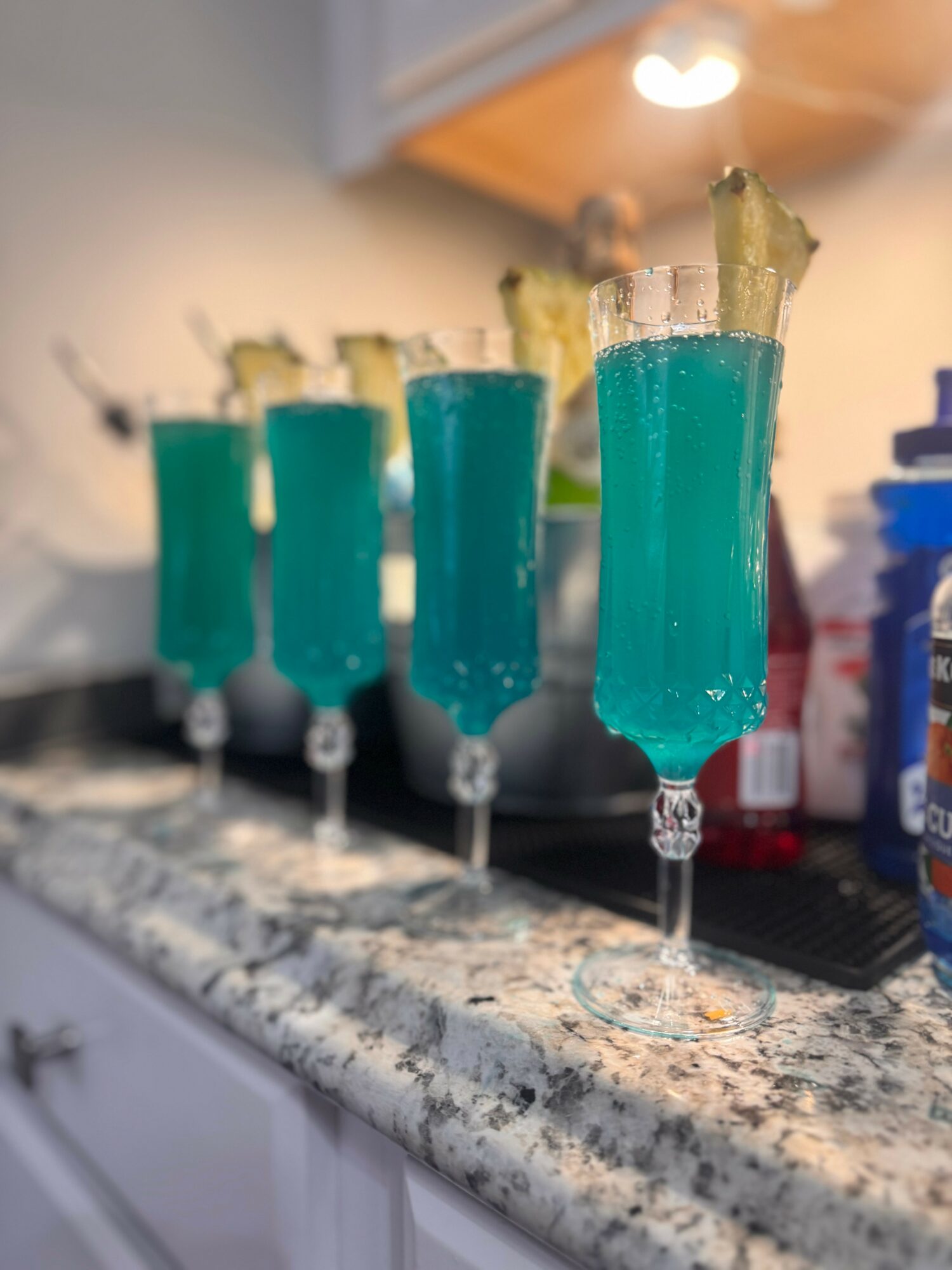 Four tall glasses with blue-green drinks and garnishes on a kitchen counter, blurred background, indoor lighting.