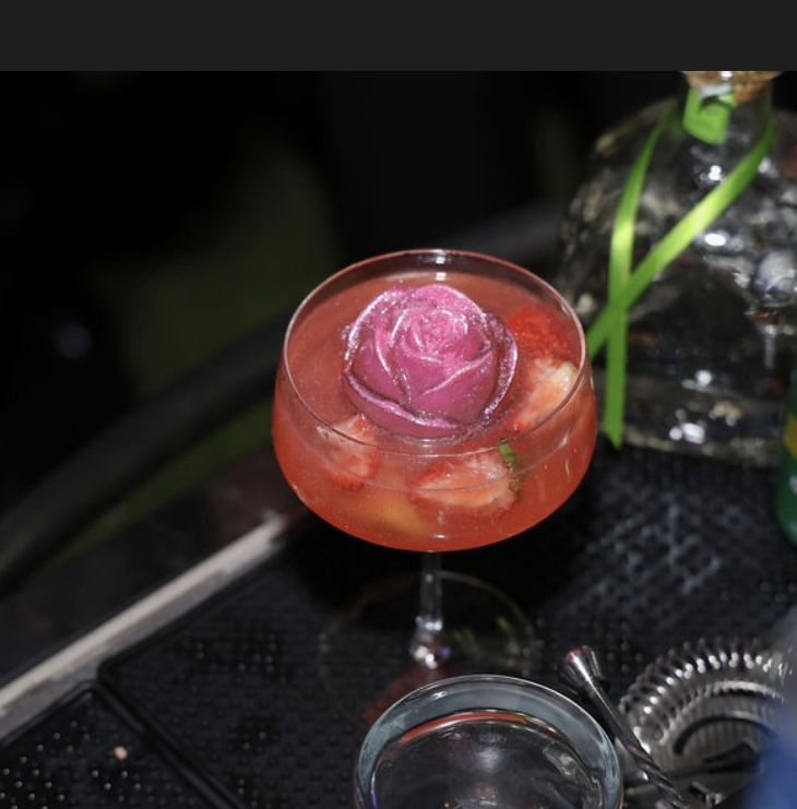 A pink flower-shaped ice cube in a pink cocktail glass with a red drink, on a black surface with a bottle in the background.