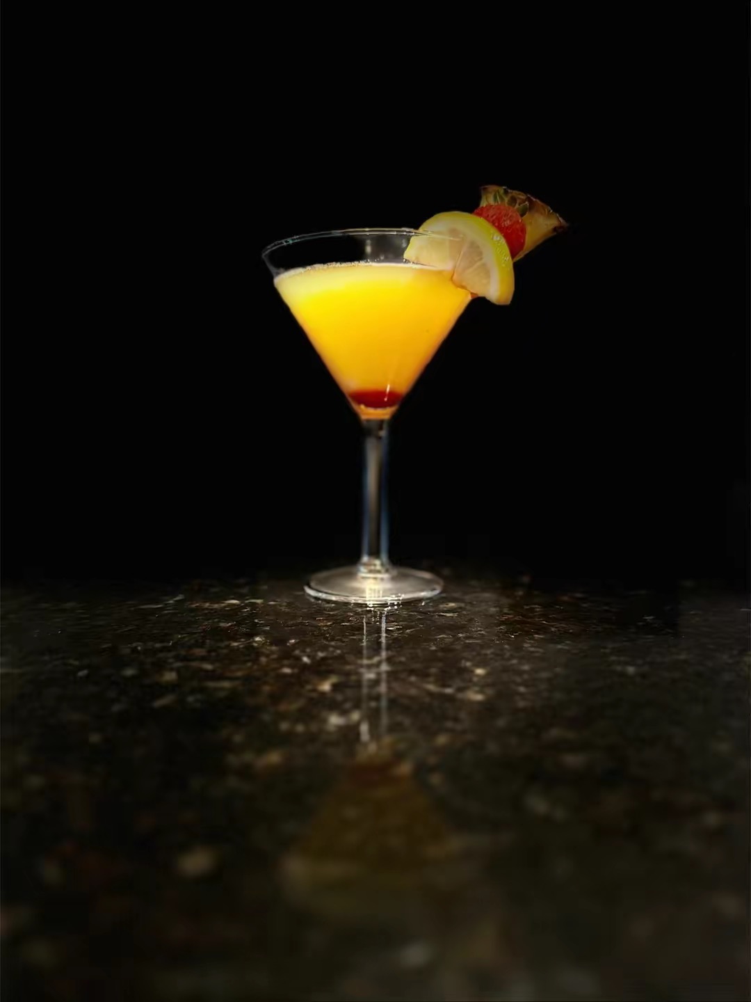 A cocktail glass with yellow drink, lemon and cherry garnishes, on a dark surface against a black background.