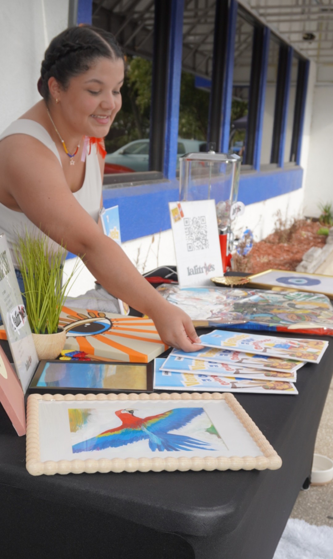 Woman smiling at a table with colorful artwork and a framed picture of a bird, outdoors near a building with blue windows.