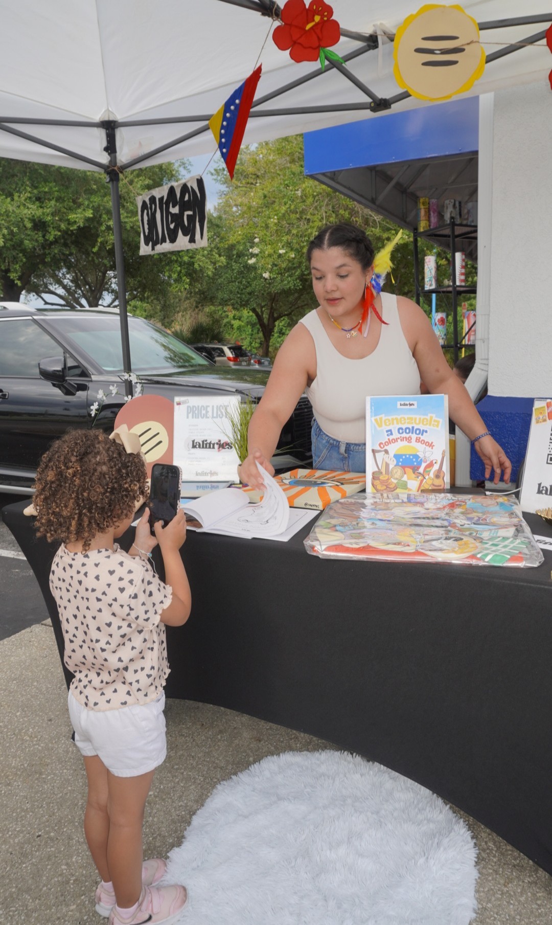 Young girl with curly hair taking a photo of a woman at an outdoor booth, with books and decorations, under a tent.