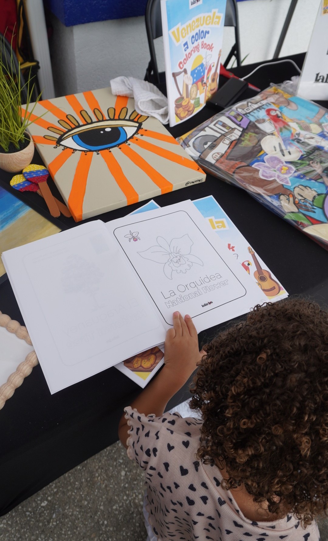 Child drawing in a notebook at a table with colorful books and a potted plant nearby.