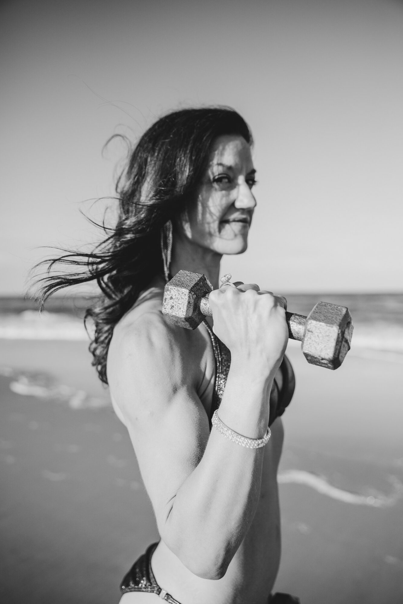 Woman holding a dumbbell on the beach, facing right, with ocean in background, black and white.