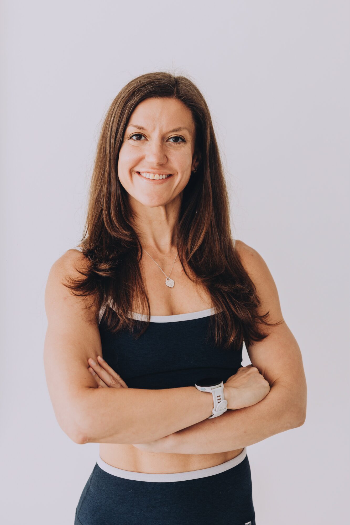 Woman with long brown hair smiling, arms crossed, wearing a black sleeveless top and a watch, standing against a plain light background.