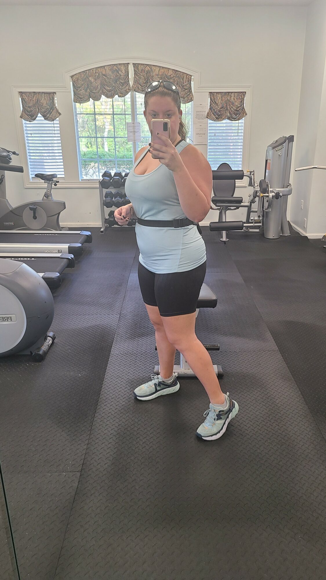 Person taking a mirror selfie in a gym, wearing a light blue top, black shorts, and sneakers, with gym equipment in background.