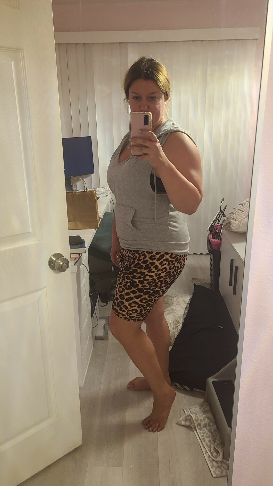 Woman taking mirror selfie in a room with light-colored walls and furniture, wearing a white top and leopard print skirt.