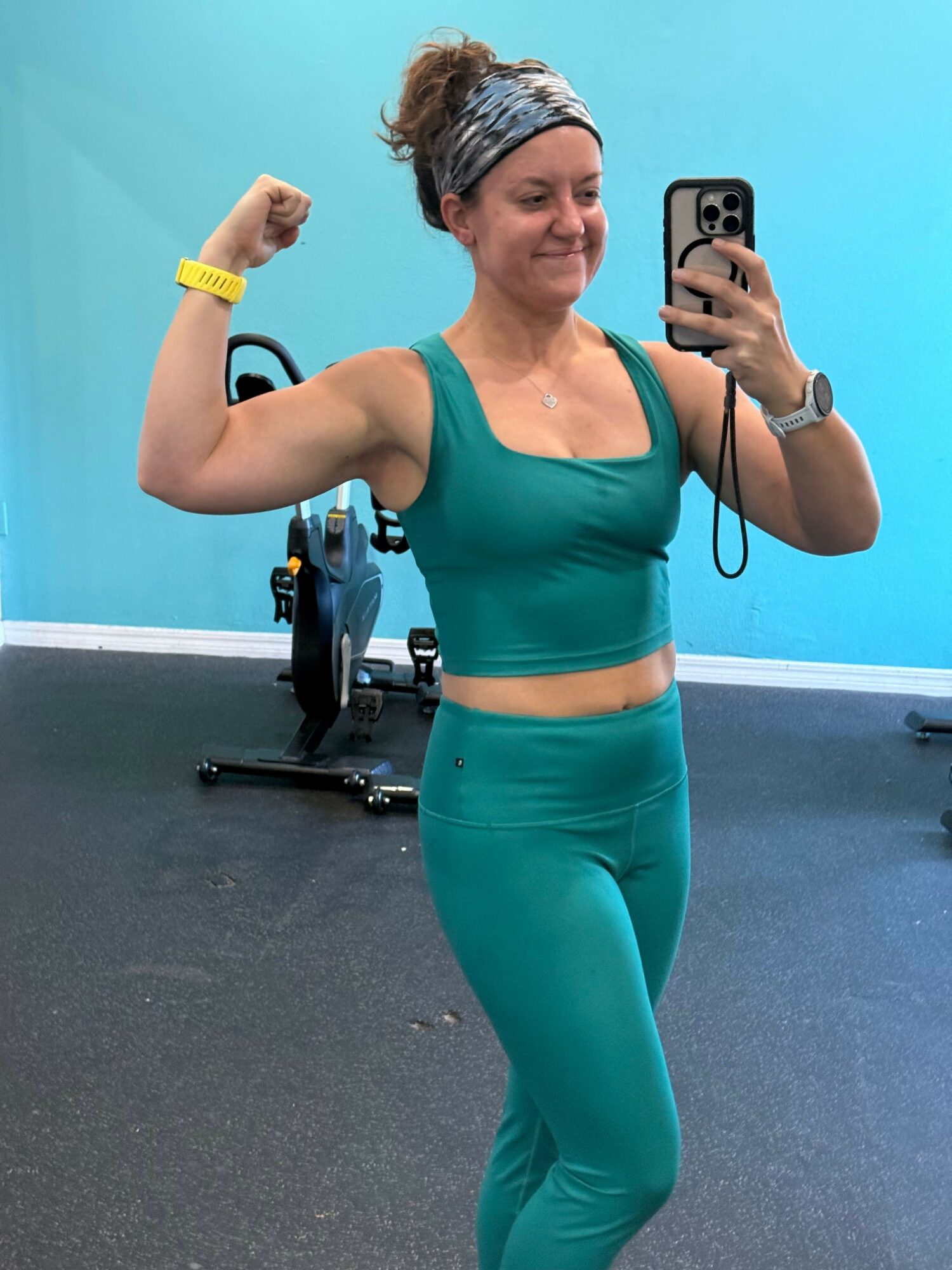 Woman in teal workout outfit flexing arm in gym, holding phone for selfie, smiling, with gym equipment in background.