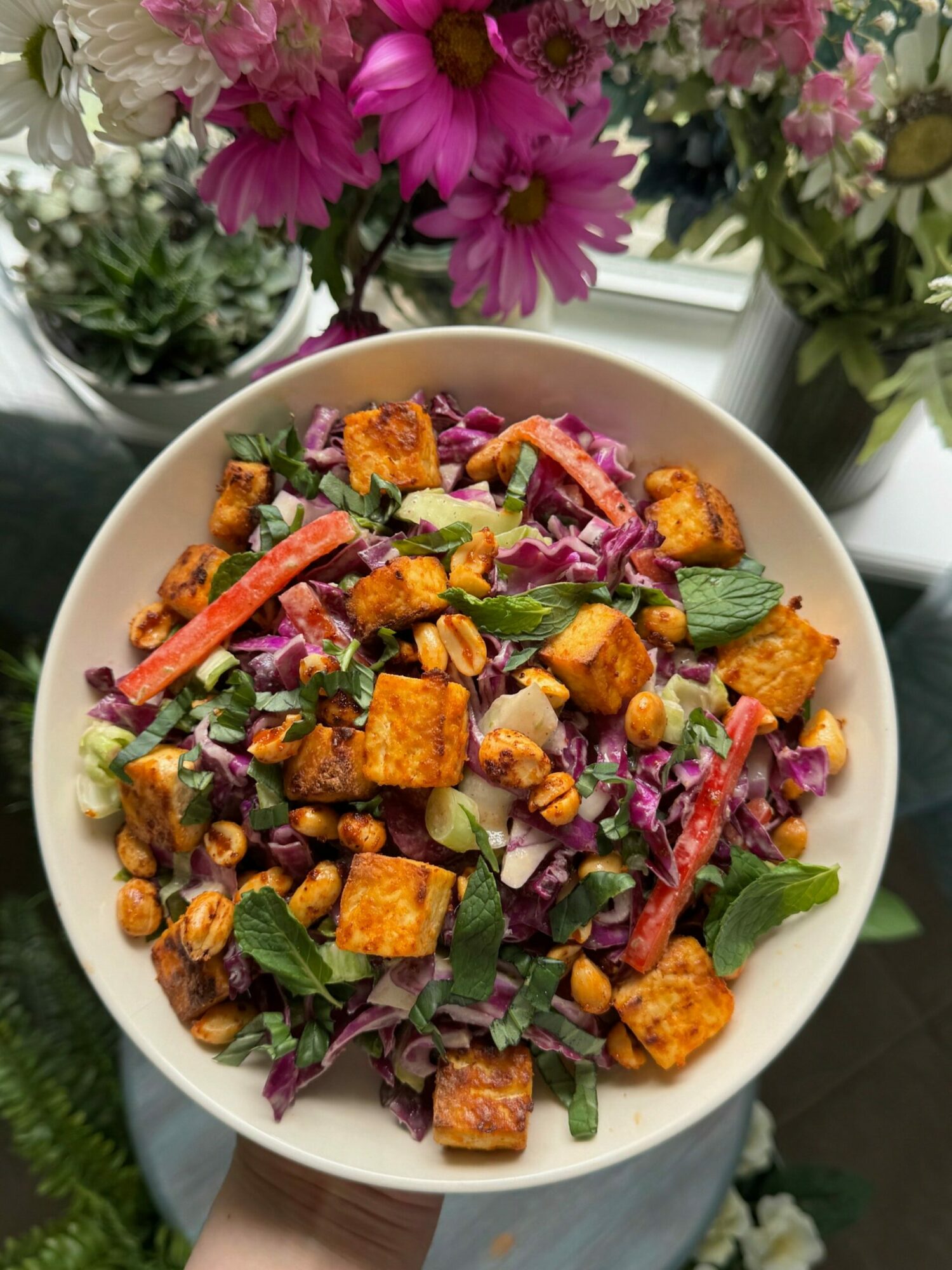Bowl of colorful salad with tofu, purple cabbage, green herbs, and red vegetables, on a reflective surface with flowers nearby.