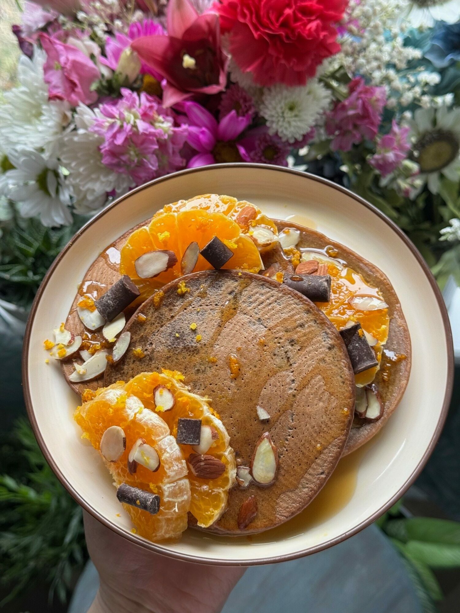 A bowl with a dessert topped with orange slices, chocolate pieces, and nuts, held by a person near colorful flowers.