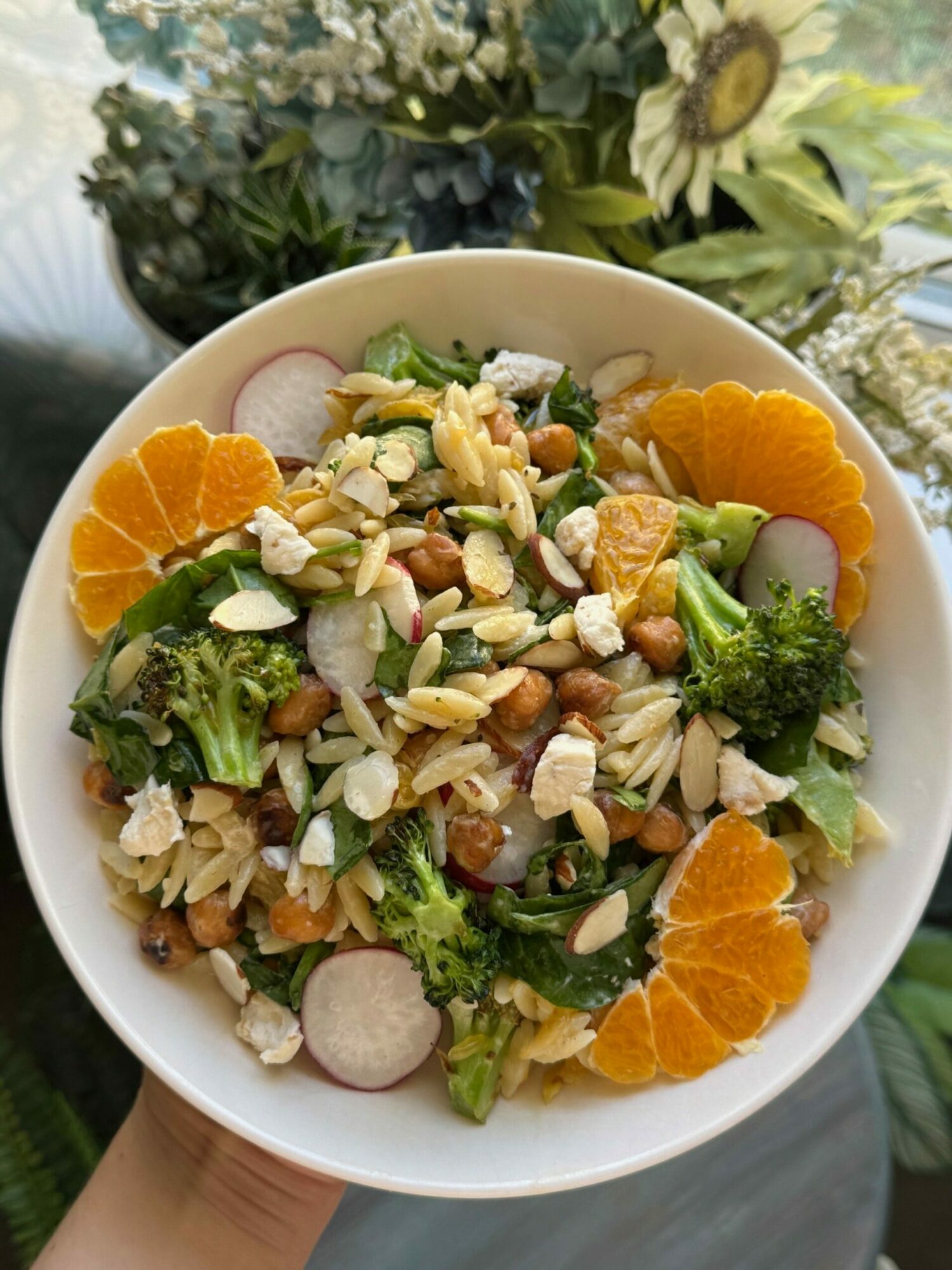 Bowl of salad with orange slices, broccoli, radish, nuts, and greens, held by a hand outdoors