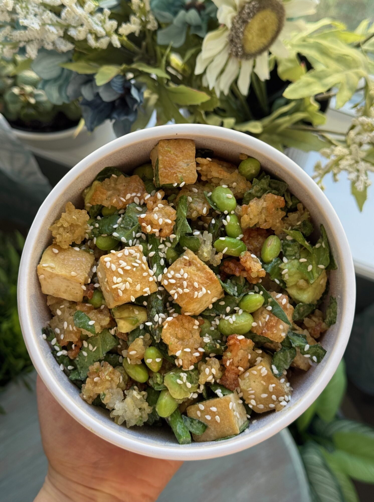 Bowl of salad with tofu, edamame, sesame seeds, and greens, held by a person, with flowers nearby.