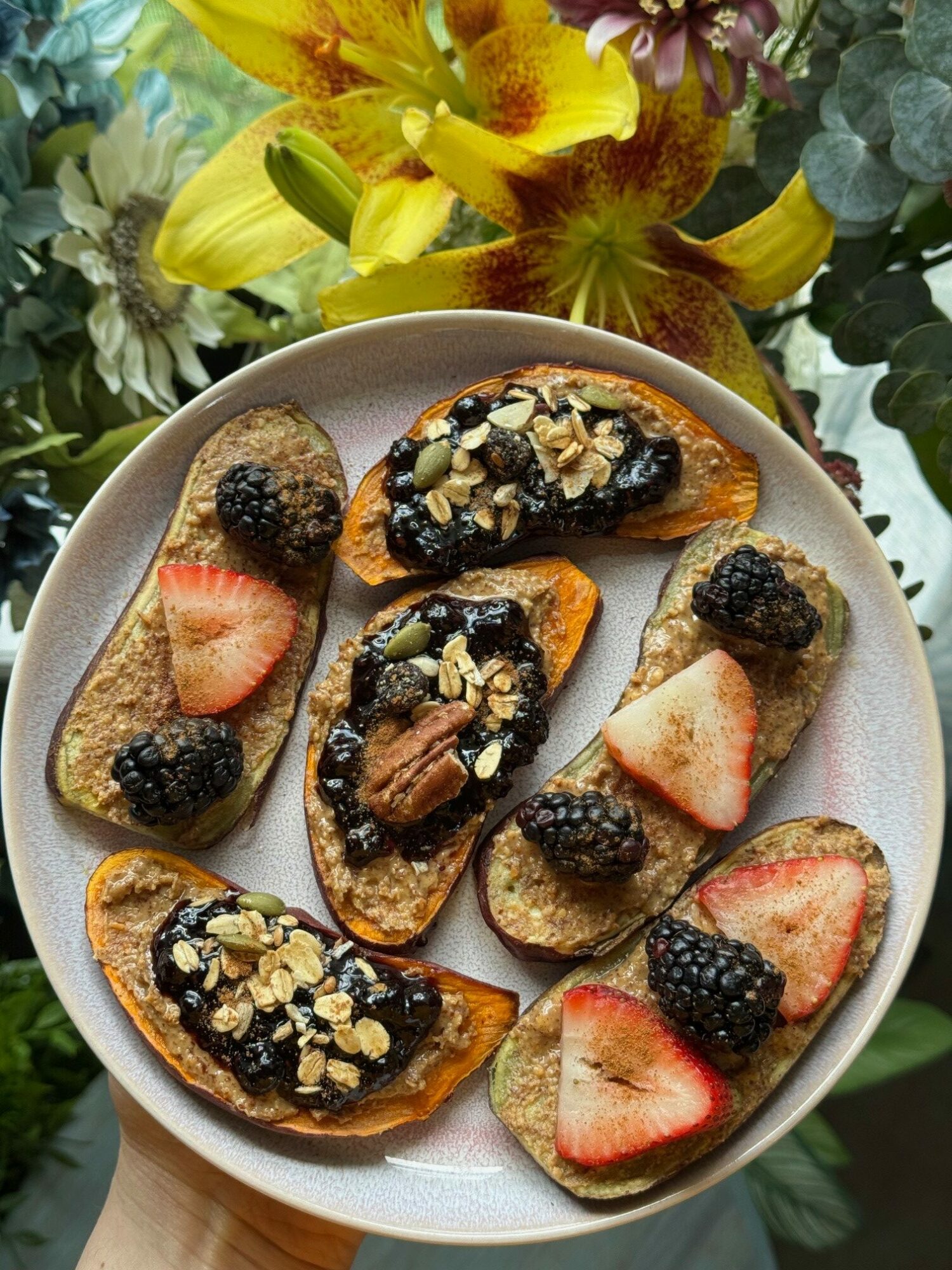 Plate with sliced strawberries, blackberries, and toasted bread topped with oats and nuts, surrounded by yellow flowers and green leaves