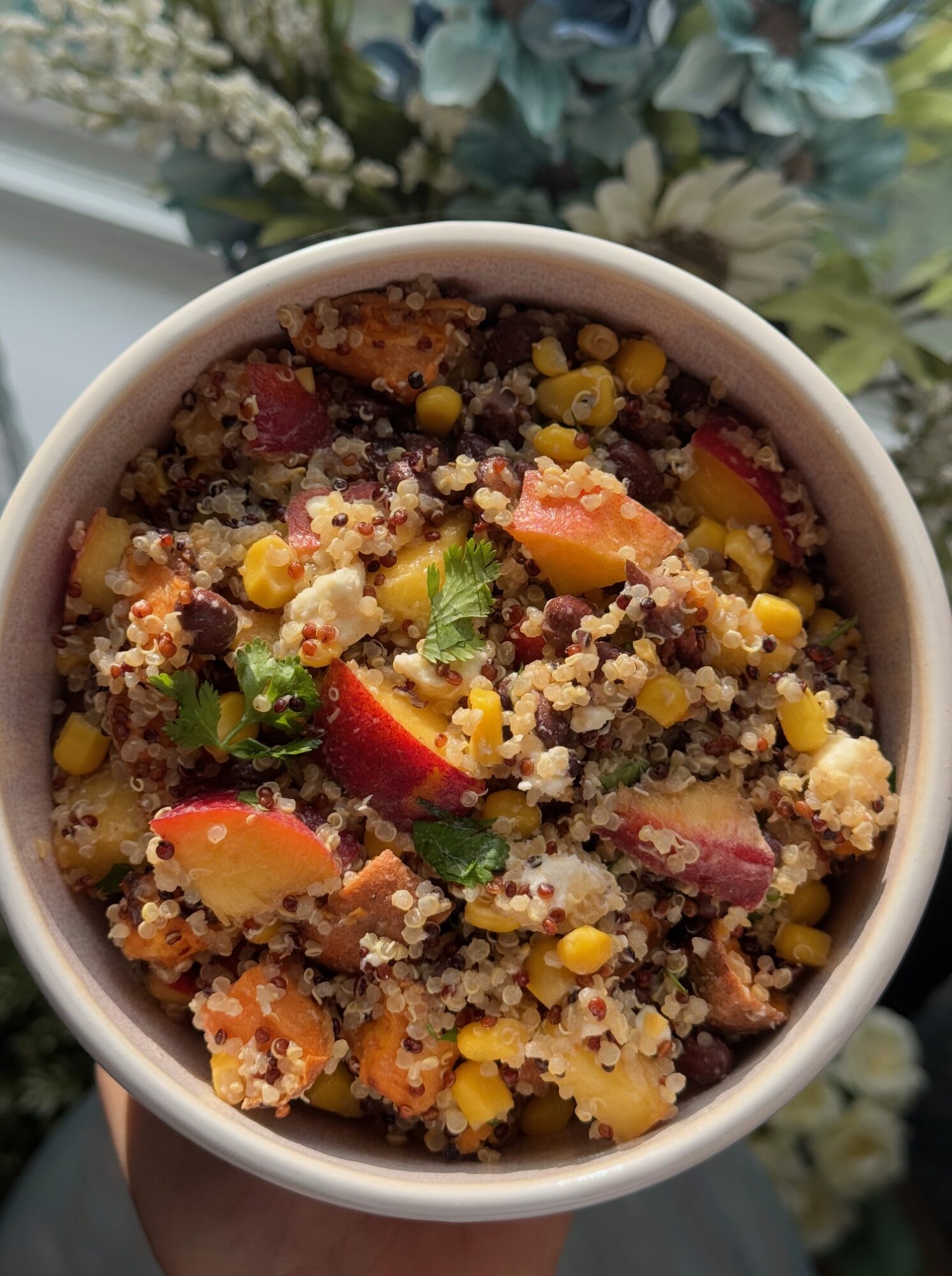 Bowl of cooked quinoa with vegetables and herbs, including corn, red bell peppers, and cilantro, on a floral background.