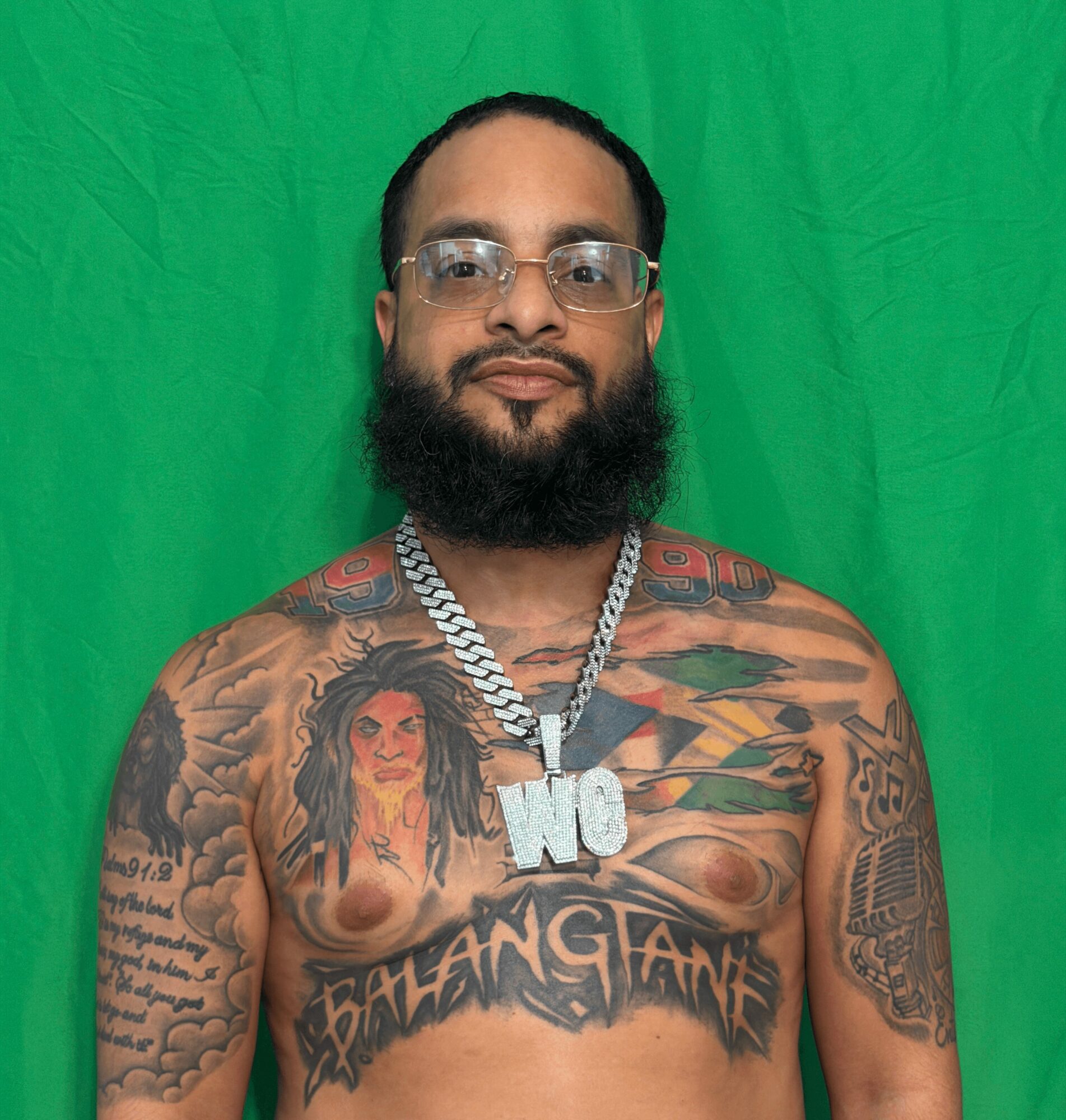 Man with glasses and a beard, shirtless, wearing a chain with a large pendant, tattoos on chest and arms, green background.