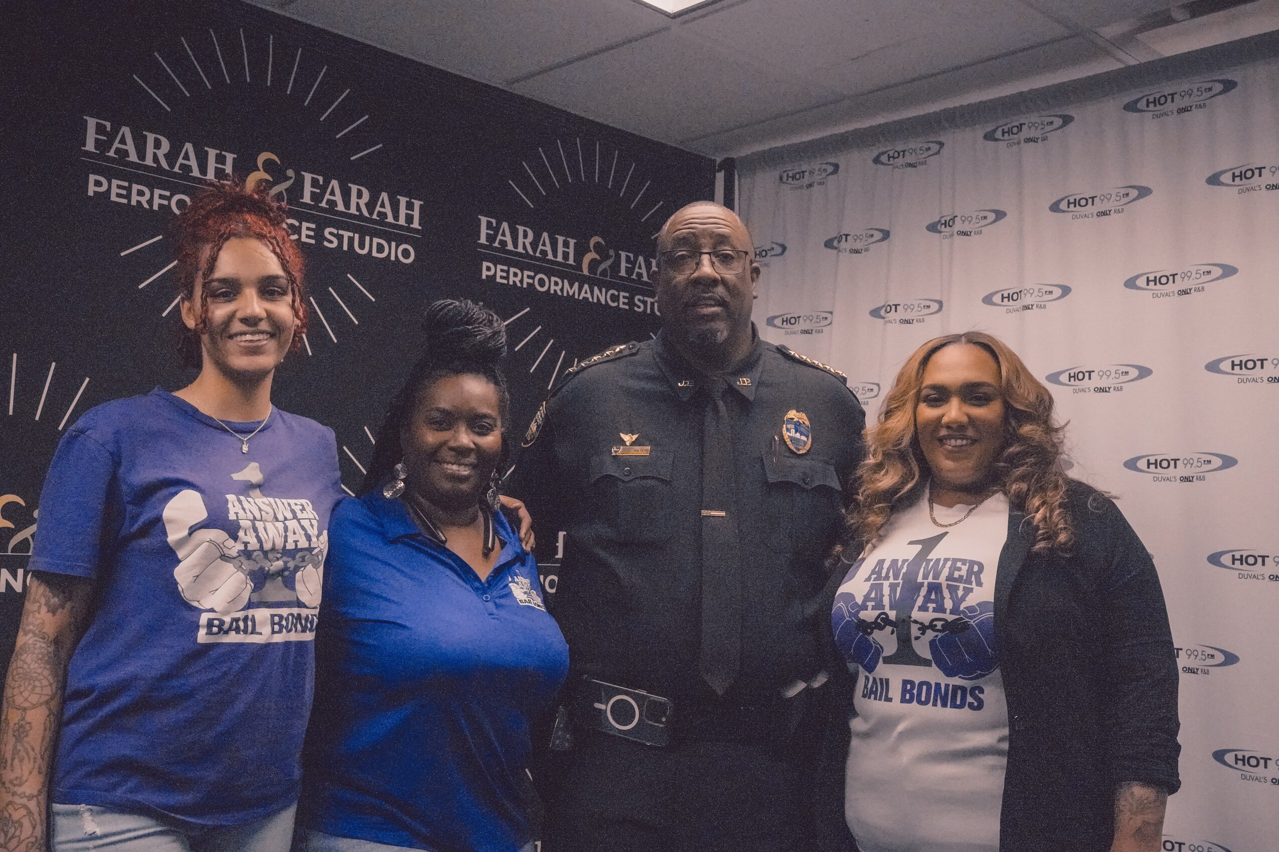 Four people standing indoors, two women wearing blue shirts, a man in a police uniform, and a woman in a white shirt and black jacket, smiling.
