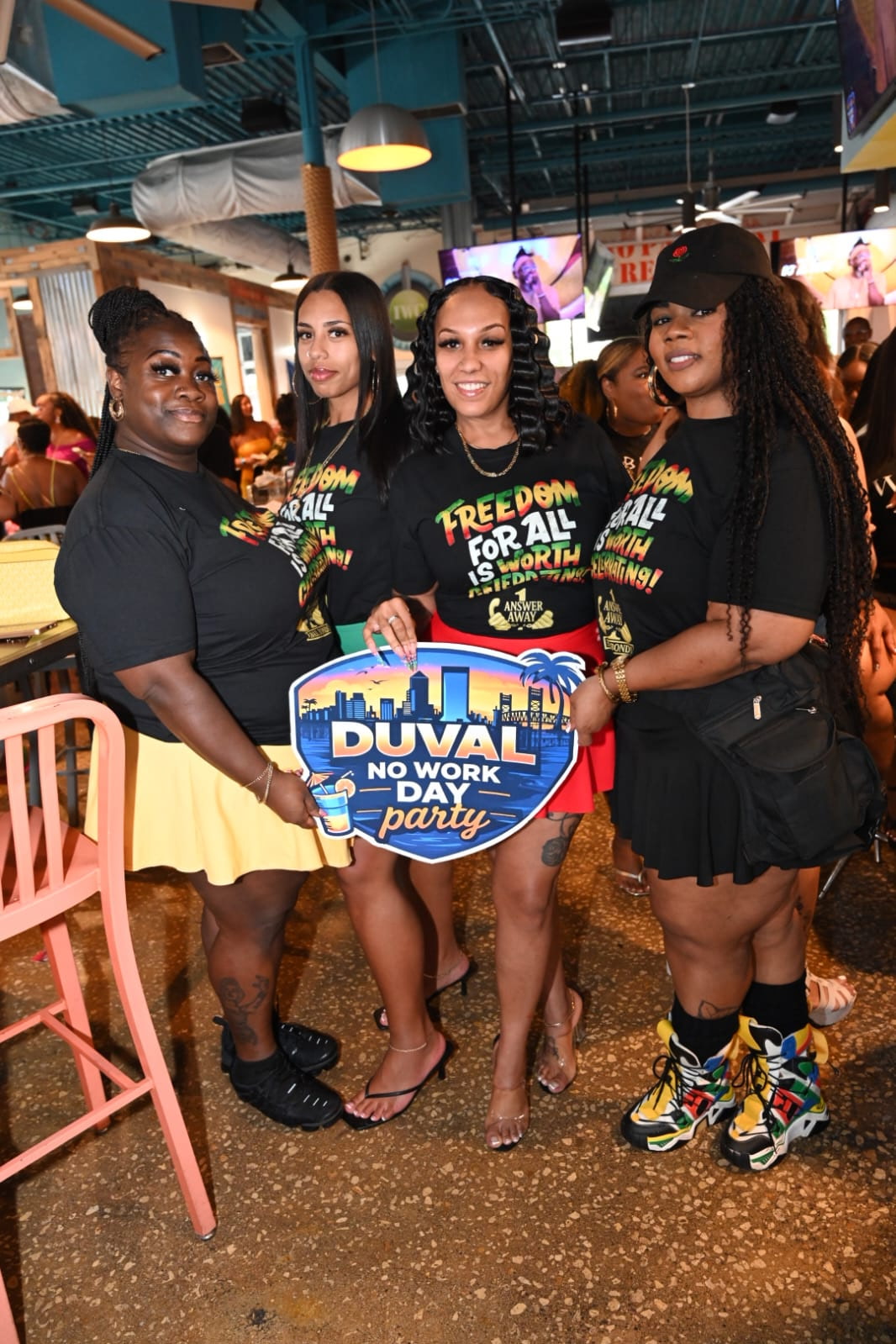 Four women holding a sign at a party, wearing matching T-shirts, in a lively indoor setting.