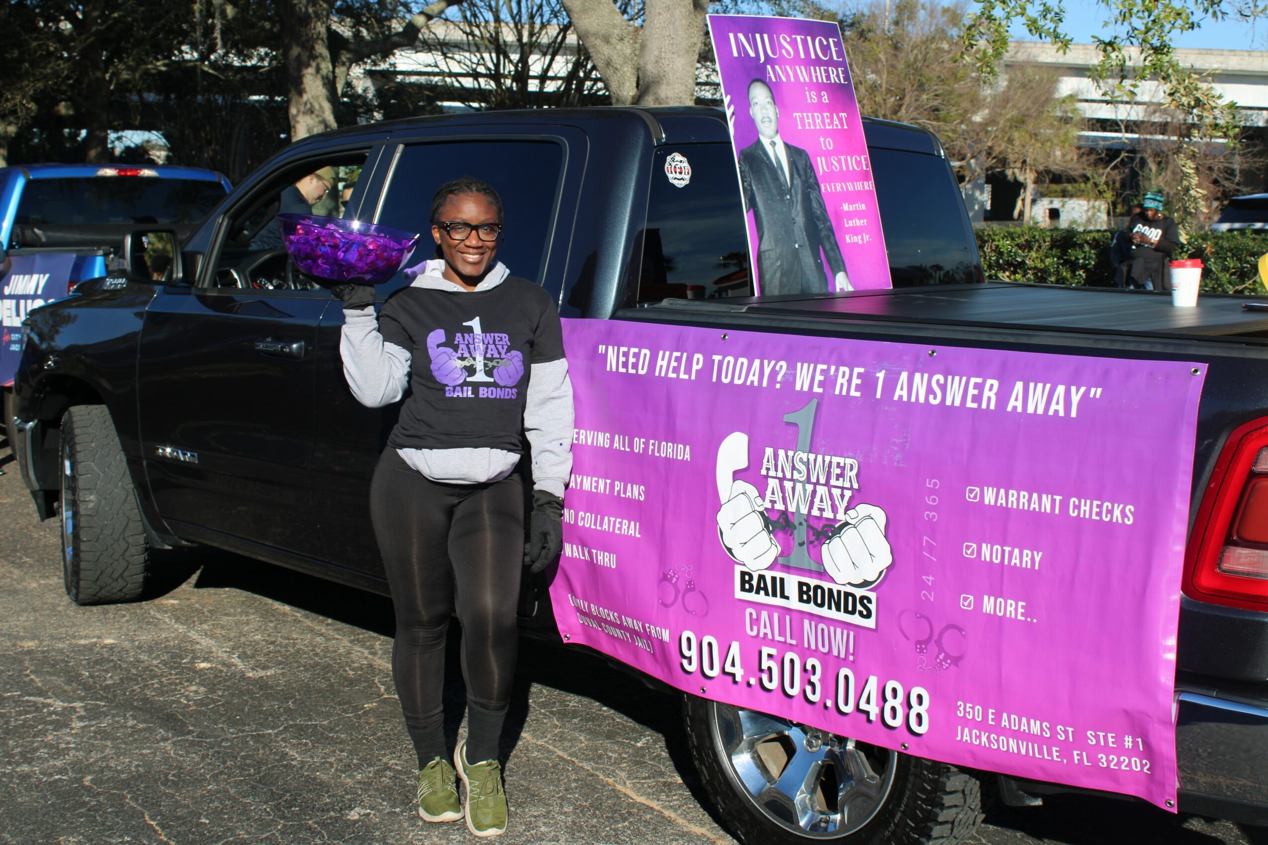 Young girl standing next to a black vehicle with purple banners and signs promoting legal aid and support services, smiling and holding a purple item.