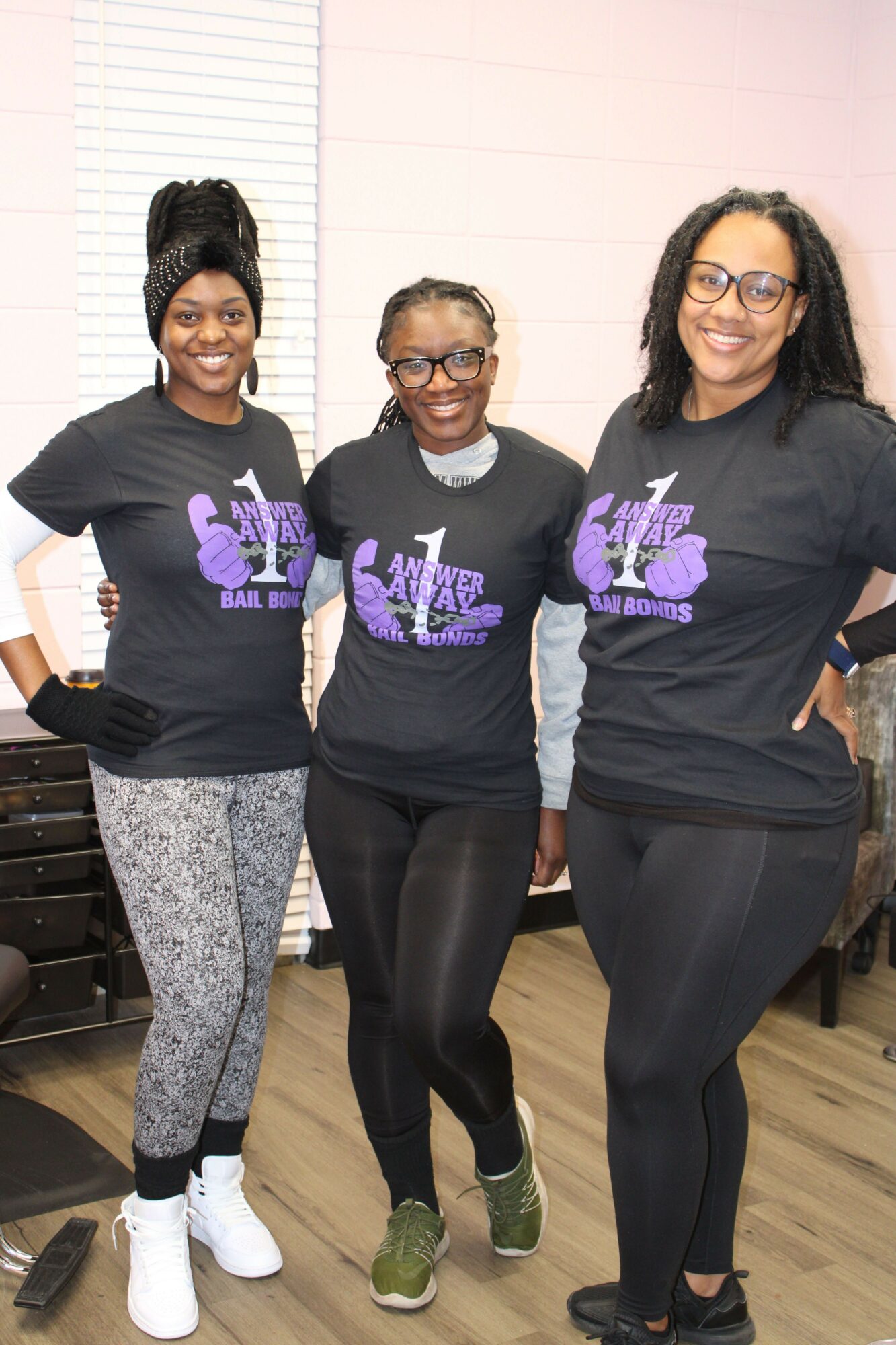 Three women standing indoors, smiling, wearing black shirts with purple designs, and casual pants or leggings.
