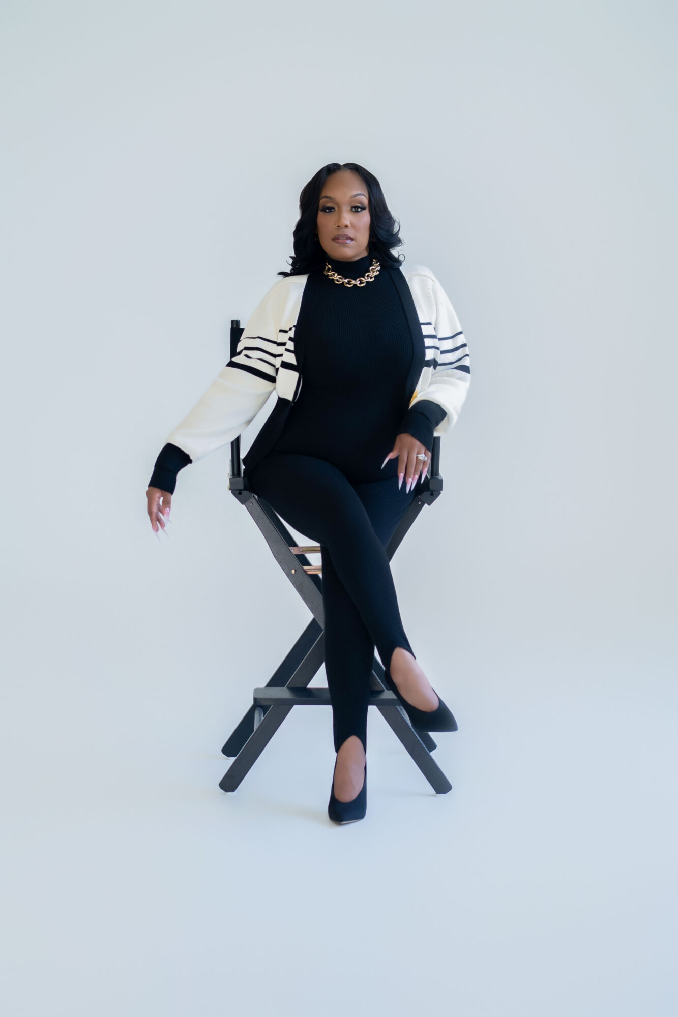 Woman sitting on a chair with crossed legs, wearing a black outfit, white jacket, and high heels, against a plain background.