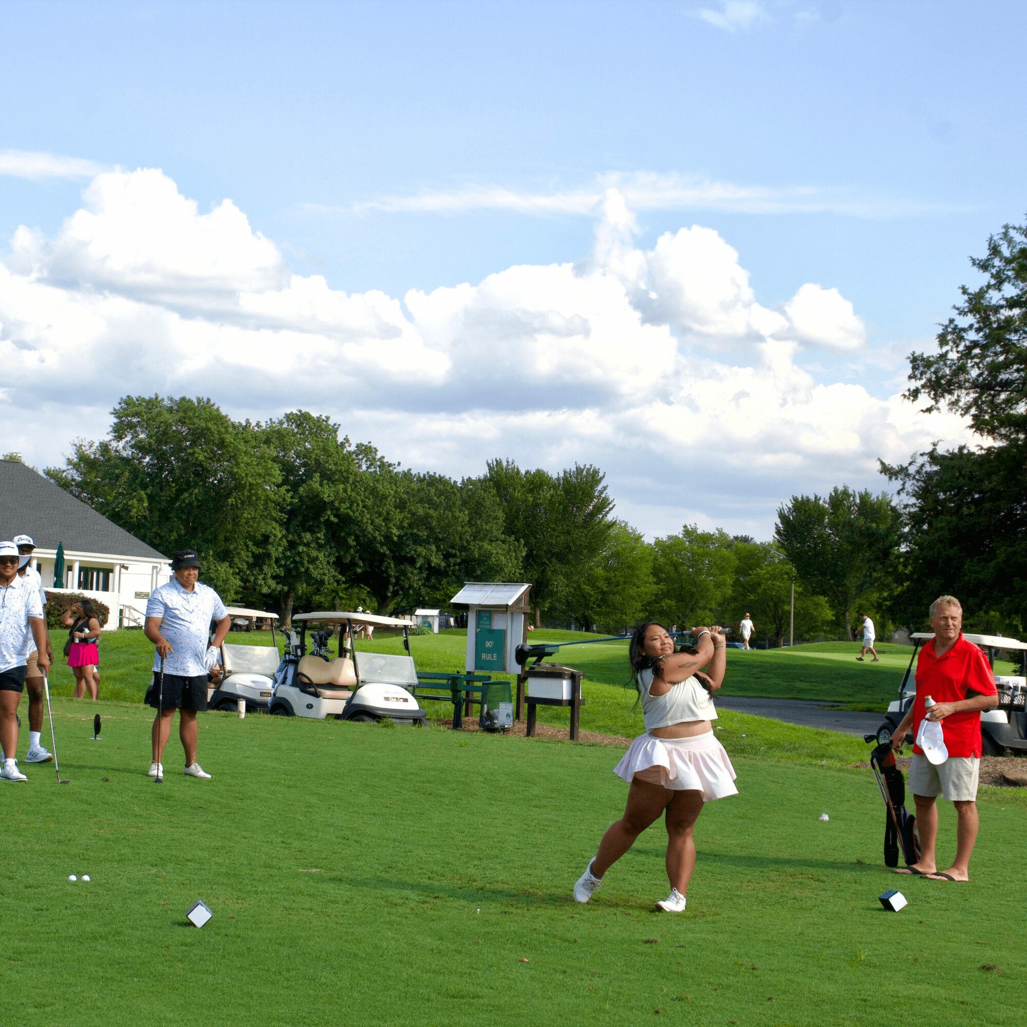 People playing golf on a grassy field with trees, cars, and a building in the background under a partly cloudy sky.