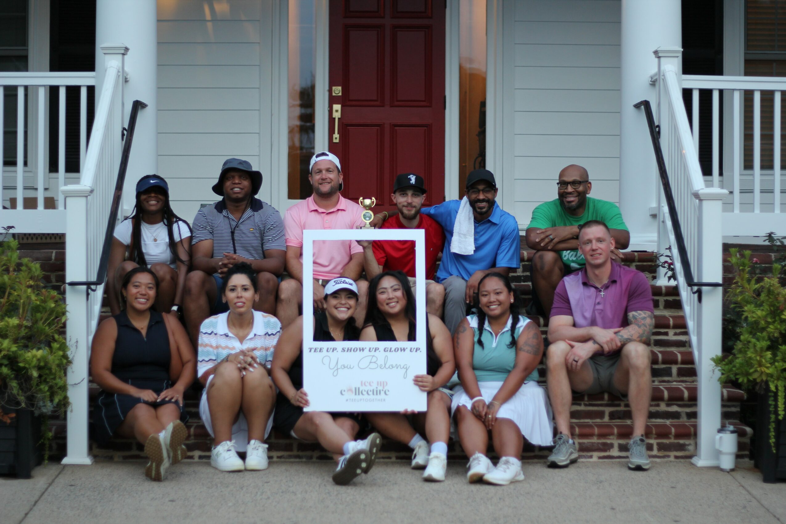 Group of ten people sitting on steps in front of a house, holding a large frame, smiling, diverse ages and styles.