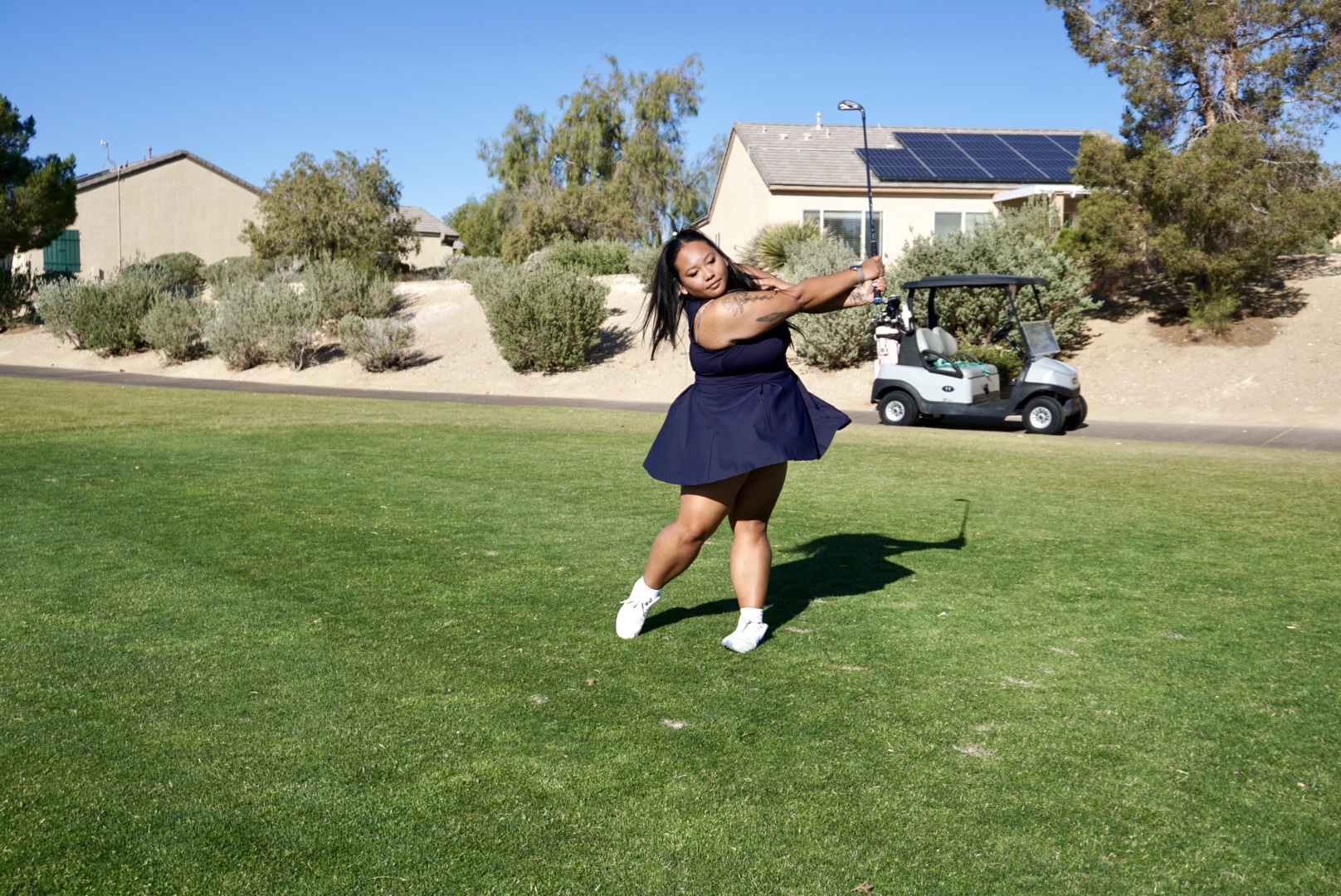 Woman in a navy dress and white shoes jumping on a grassy field with houses and trees in the background.
