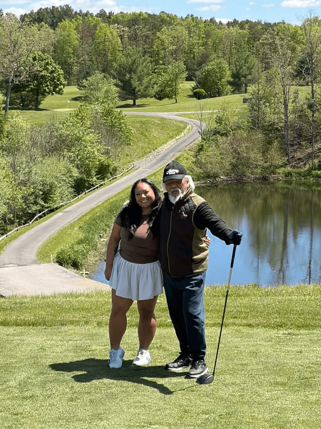Two people stand on grass near a pond with trees and a winding path in the background, smiling at the camera.