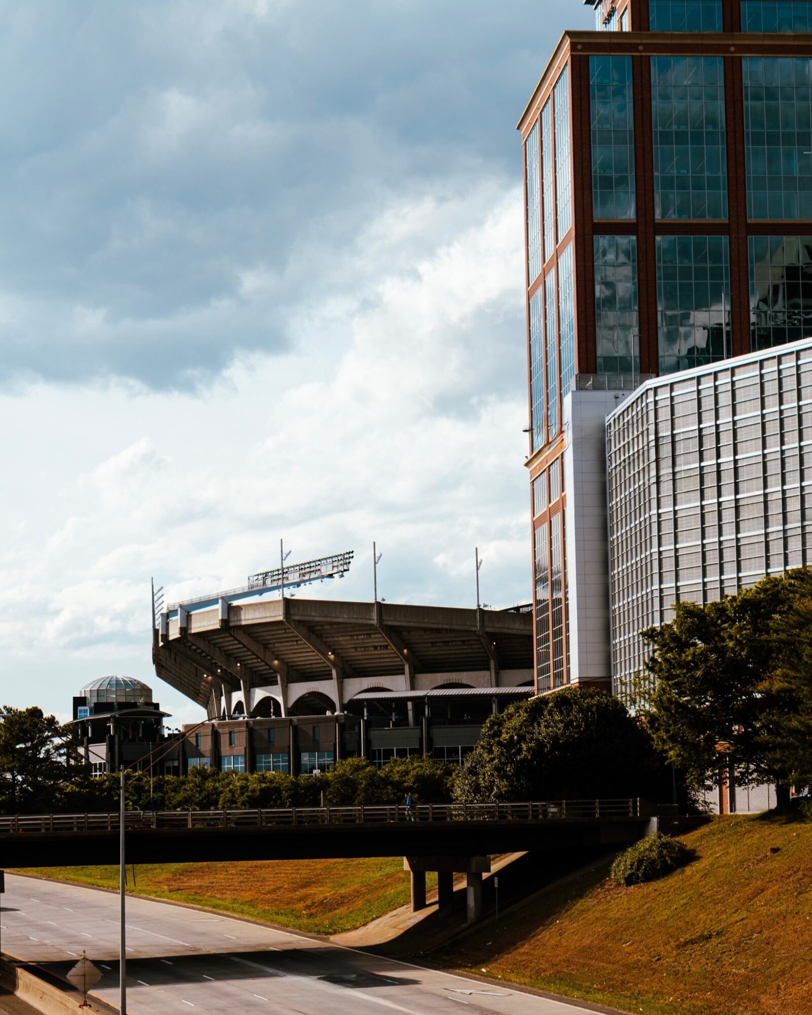Modern glass building with a stadium and cloudy sky in the background, trees and a road in the foreground.